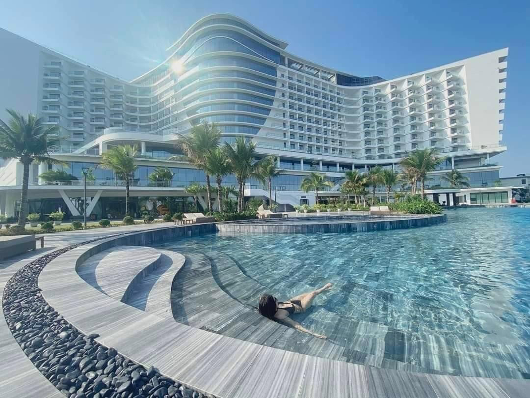 Woman relaxing in a pool with a large modern hotel and palm trees in the background on a sunny day.