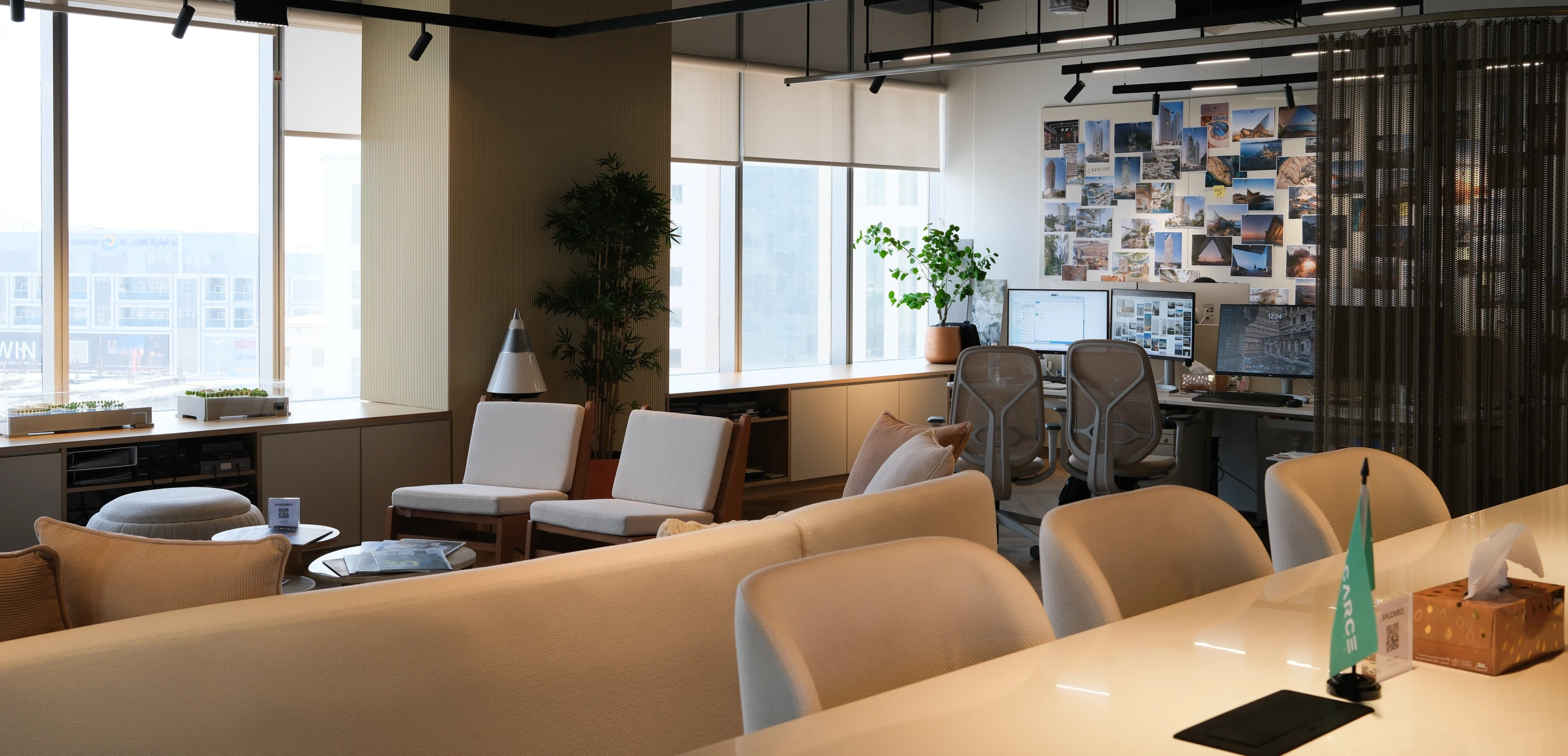 Modern office lounge and workspace with beige chairs, sofas, plants, and dual computer monitors.