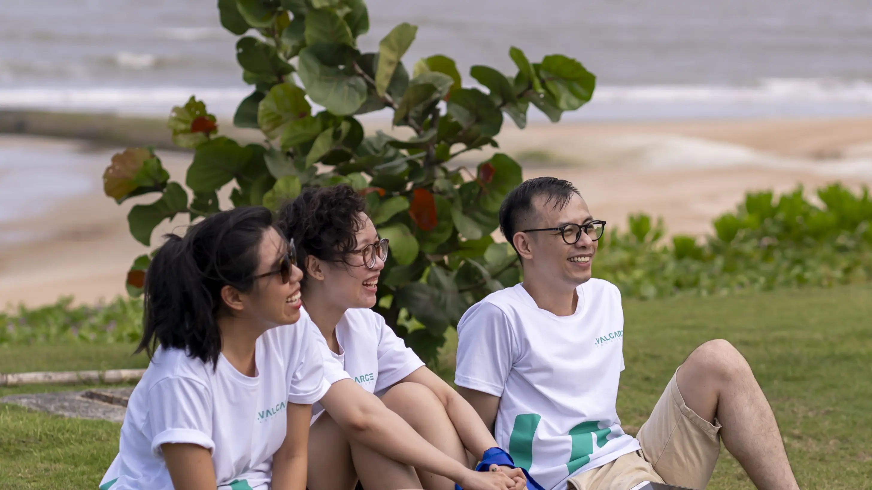 Three young people sitting on grass by the beach, smiling and enjoying a sunny day.
