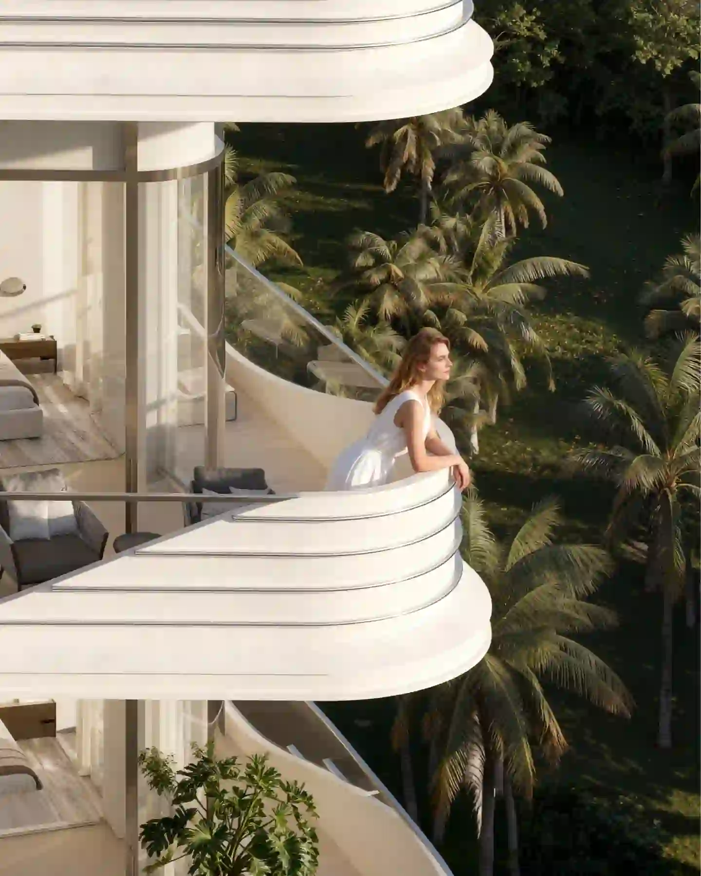 Woman in white dress leaning on balcony railing overlooking tropical palm trees and greenery.