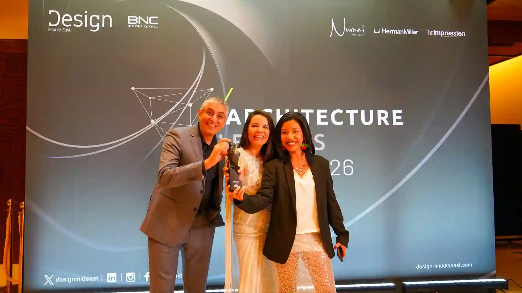 Three people smiling at an award event with a trophy in front of a Design Middle East backdrop.