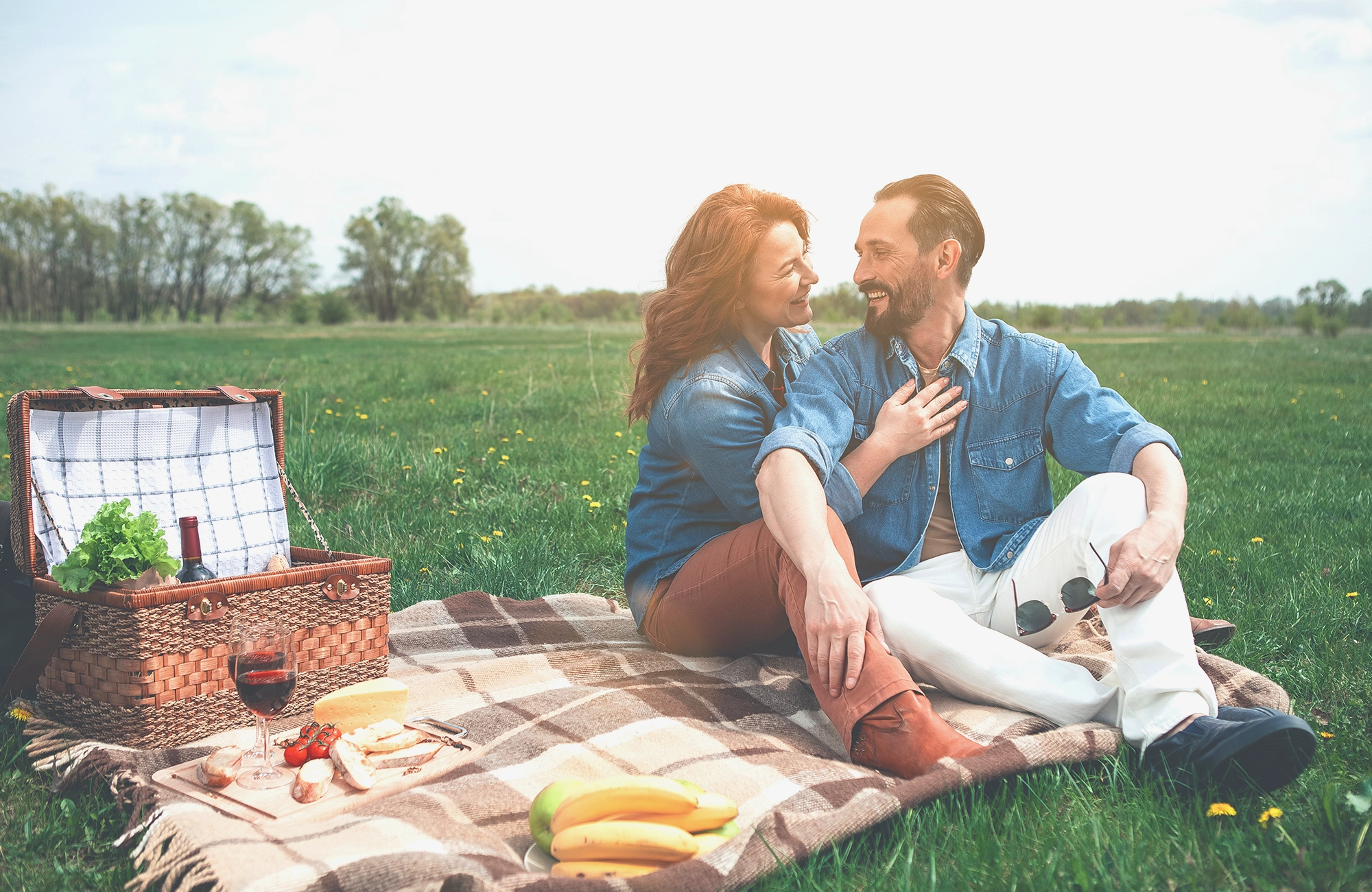 Paar sitzt auf kariertem Picknickdecke auf grüner Wiese mit Picknickkorb, Wein und Früchten.
