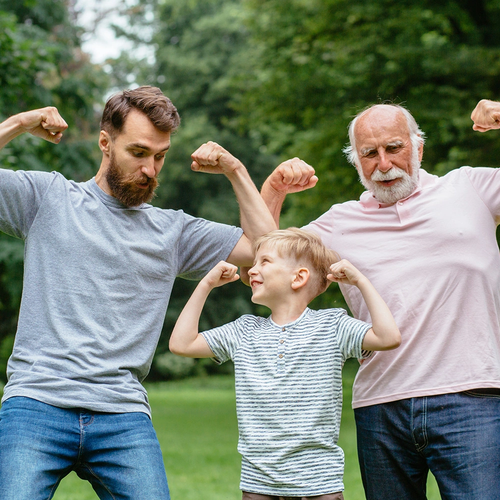 Drei Generationen – ein Kind, ein Mann und ein älterer Mann – zeigen im Park ihre Muskeln und lächeln.