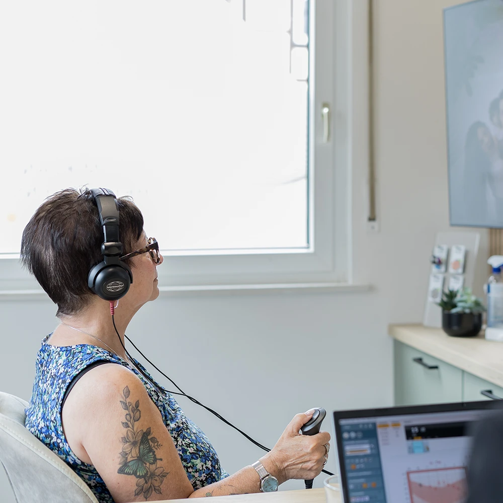 Frau mit Kopfhörern und Tattoo am Arm testet Hörgerät in moderner Praxis.