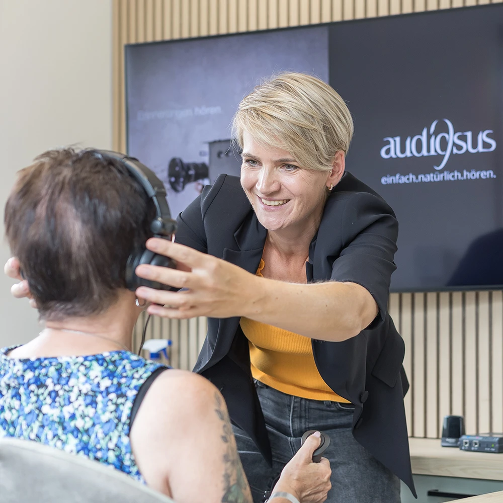 Frau setzt einem Kunden Kopfhörer auf, im Hintergrund Bildschirm mit Logo 'audiosus'.