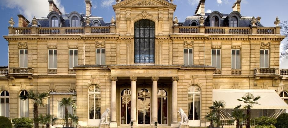 Façade imposante d’un bâtiment classique en pierre avec colonnes, statues de lions, et palmiers.