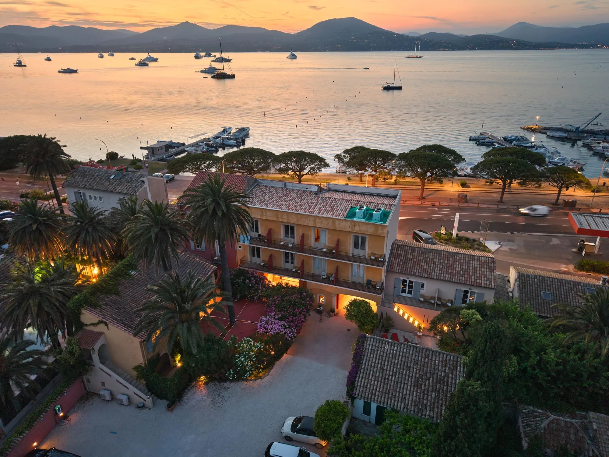 Aerial view of a waterfront hotel at sunset with boats and mountains in the background.
