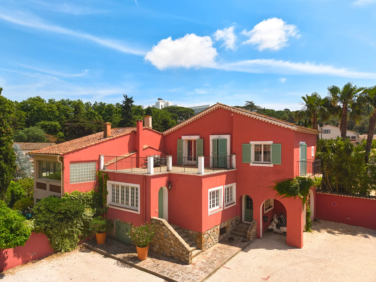 Villa rouge avec balcons, volets verts et plantes, sous un ciel bleu avec quelques nuages.