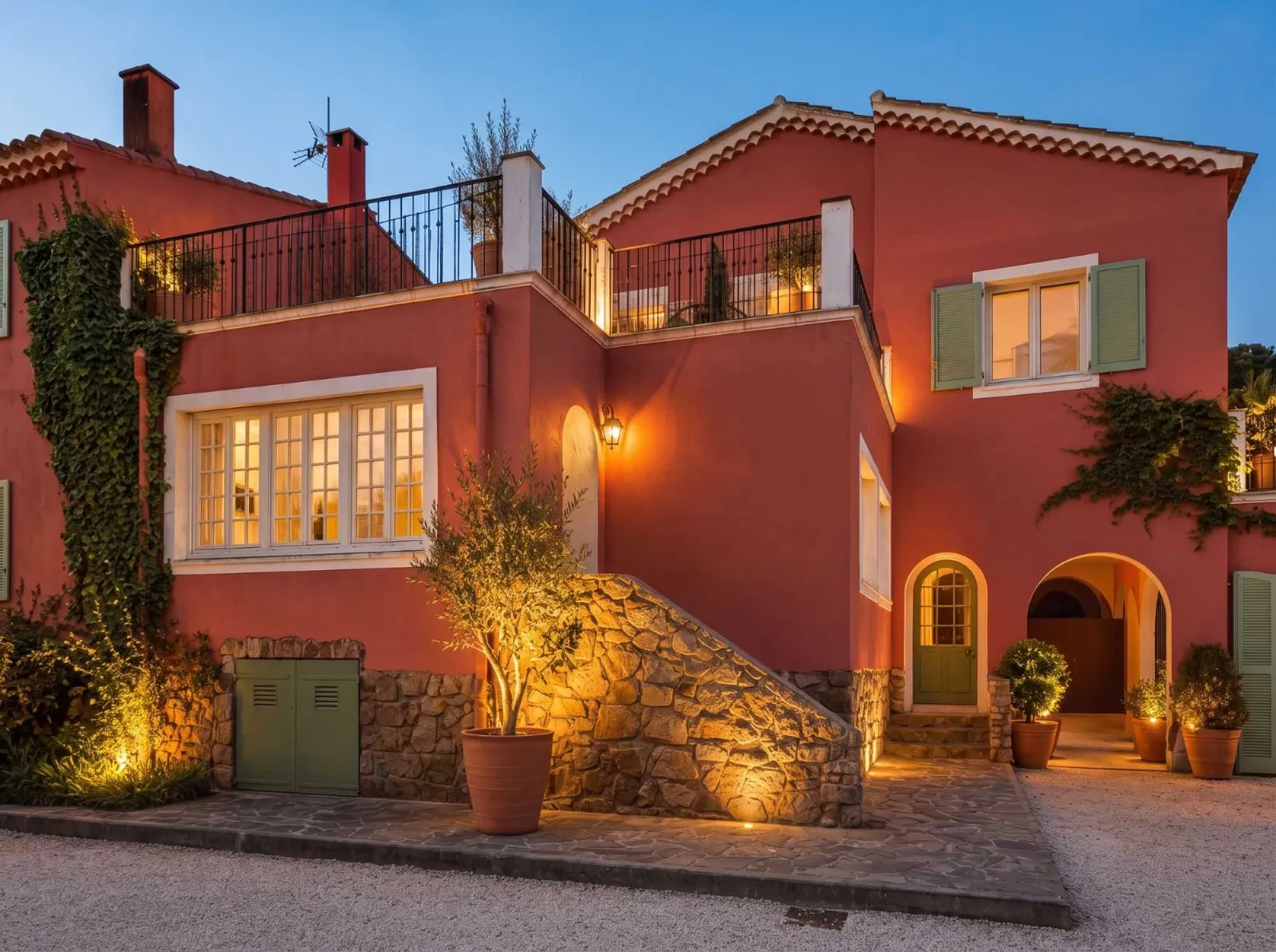 Grande villa rose avec toits en tuiles, volets verts, terrasse et végétation sous ciel bleu clair.