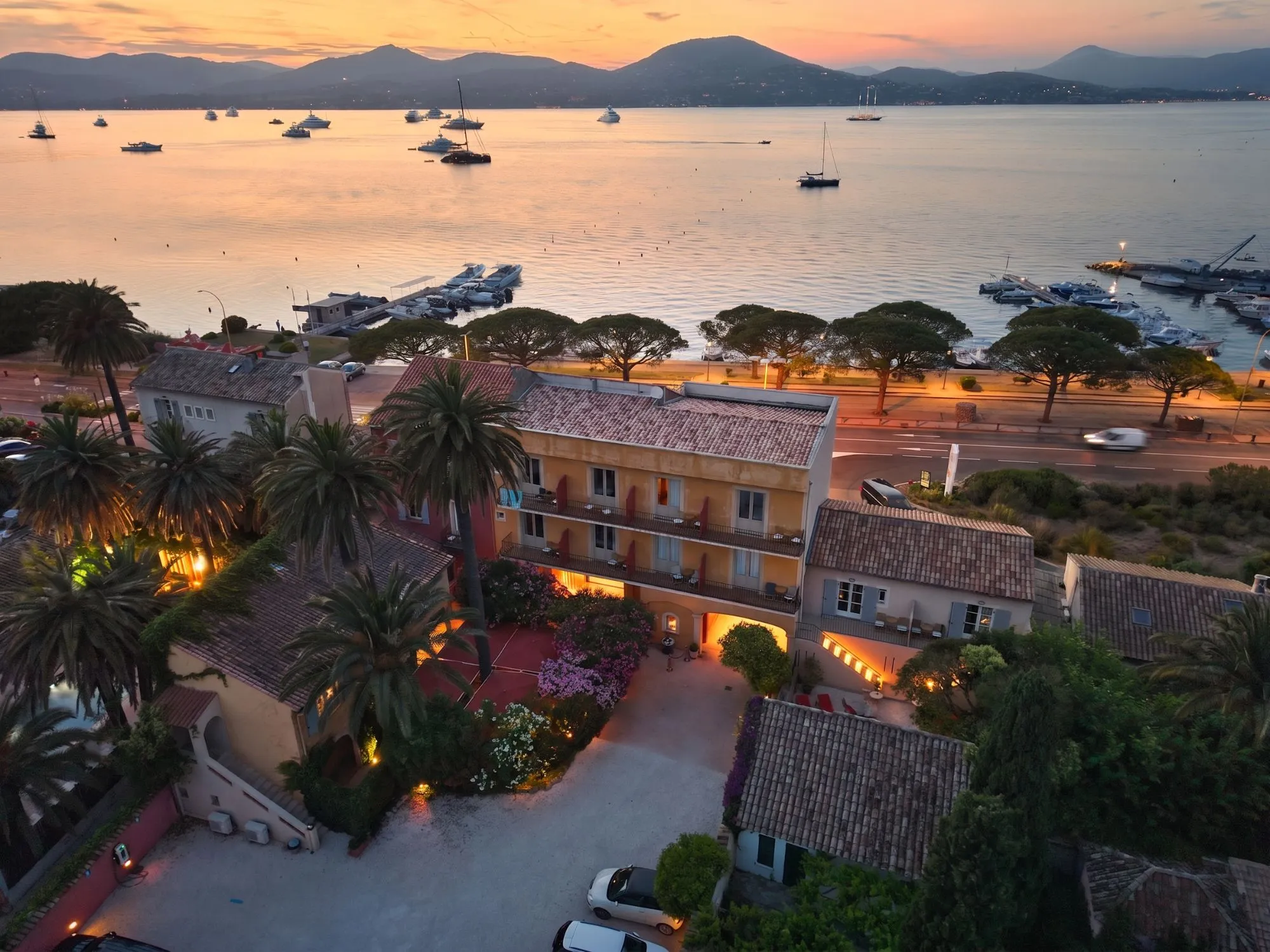 Vue aérienne d'un hôtel au bord de l'eau au coucher du soleil avec des bateaux et des montagnes en arrière-plan.