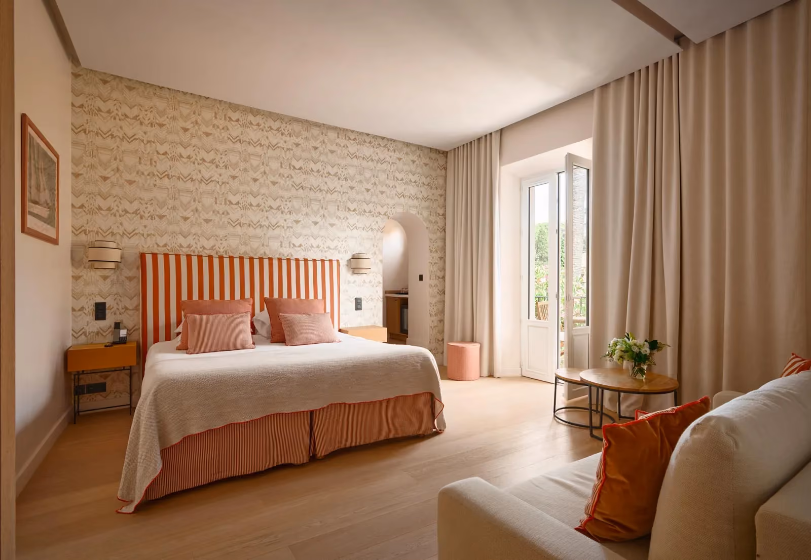 Chambre d'hôtel lumineuse avec lit à tête de lit rayée orange et blanc, coussins roses, papier peint à motifs beige, canapé et balcon ouvrant sur la verdure.