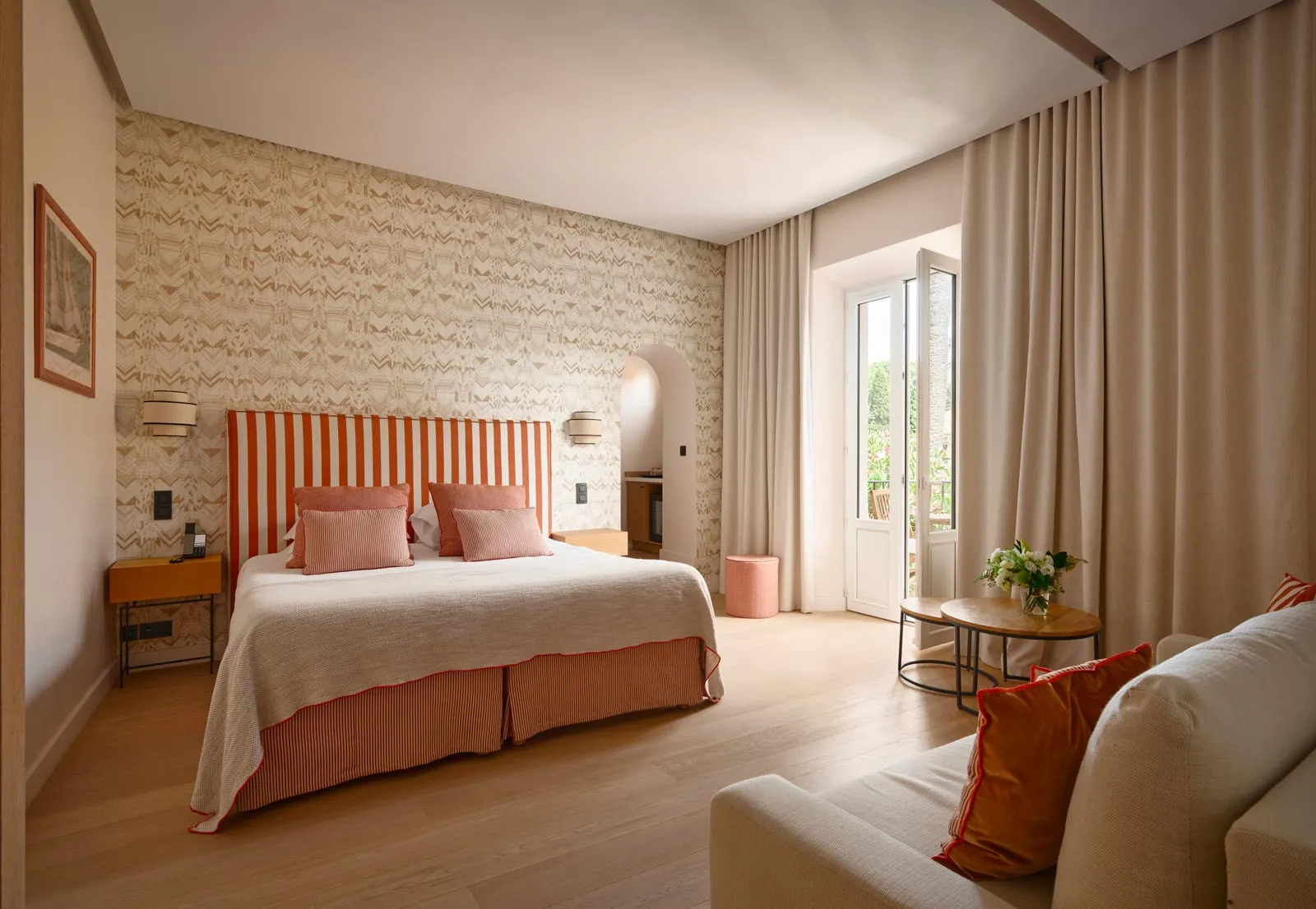 Camera d'albergo luminosa con letto dotato di testiera a righe arancioni e bianche, cuscini rosa, carta da parati con motivi beige, divano e balcone affacciato sul verde.