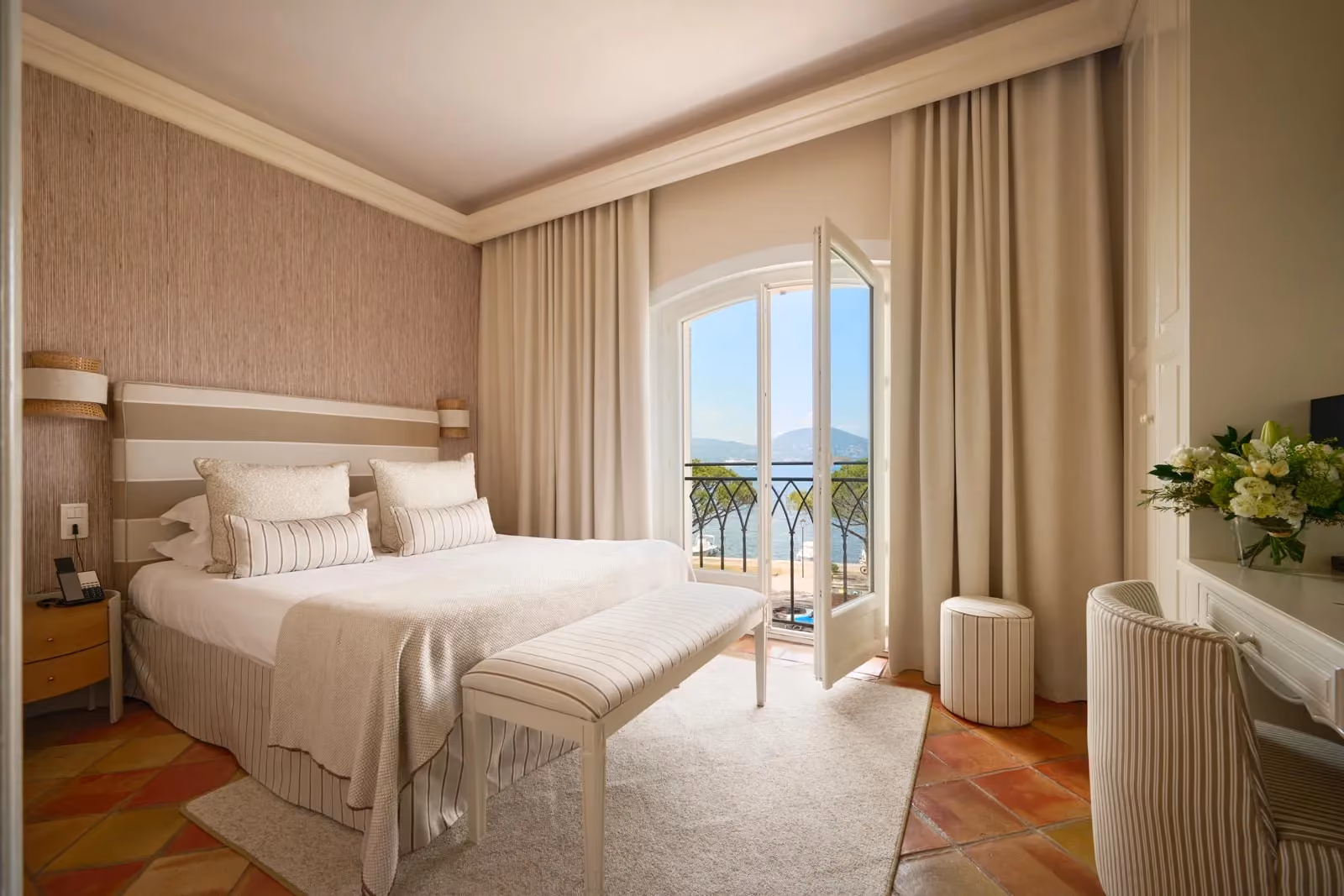 Chambre d'hôtel élégante aux tons beige et blanc, avec lit rayé, sol en terre cuite, et porte-fenêtre ouverte sur un balcon en fer forgé donnant sur la mer et les collines.