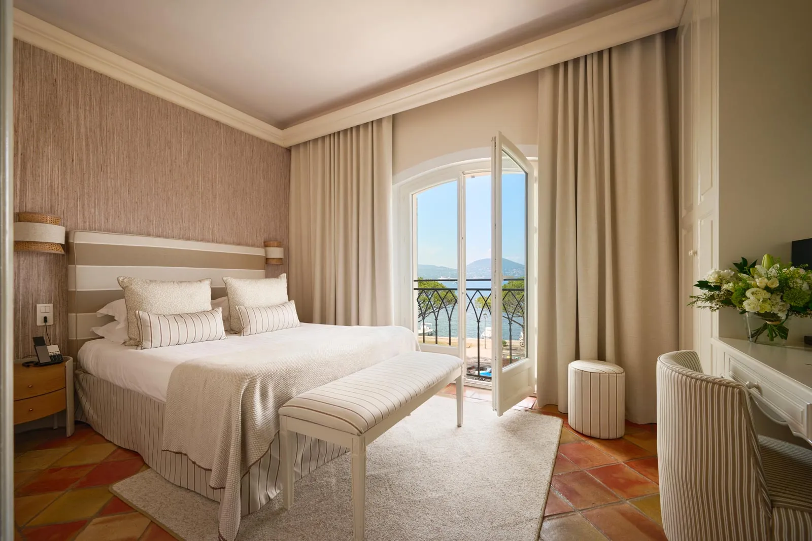 Elegante camera d'albergo nei toni del beige e del bianco, con letto a righe, pavimento in cotto e porta-finestra aperta su un balcone in ferro battuto affacciato sul mare e sulle colline.