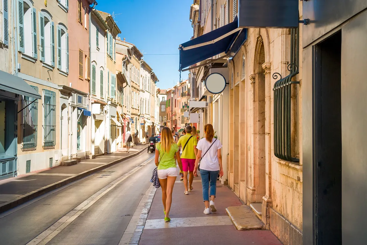 Rue commerçante ensoleillée bordée de bâtiments pastel aux volets colorés, avec des passants flânant devant des boutiques à Saint-Tropez.