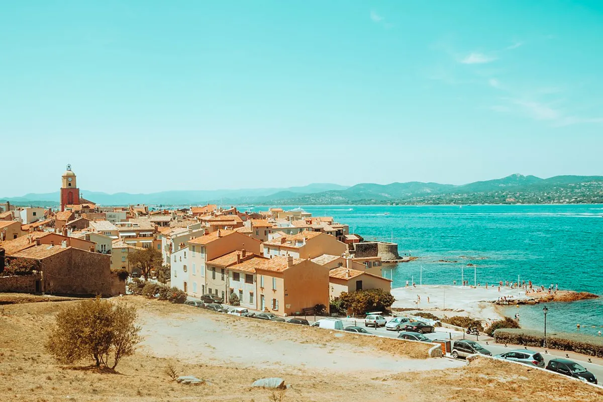 Vue panoramique de Saint-Tropez avec son clocher rouge, ses maisons aux toits de tuiles bordant la plage, et la mer turquoise s'étendant vers les collines.