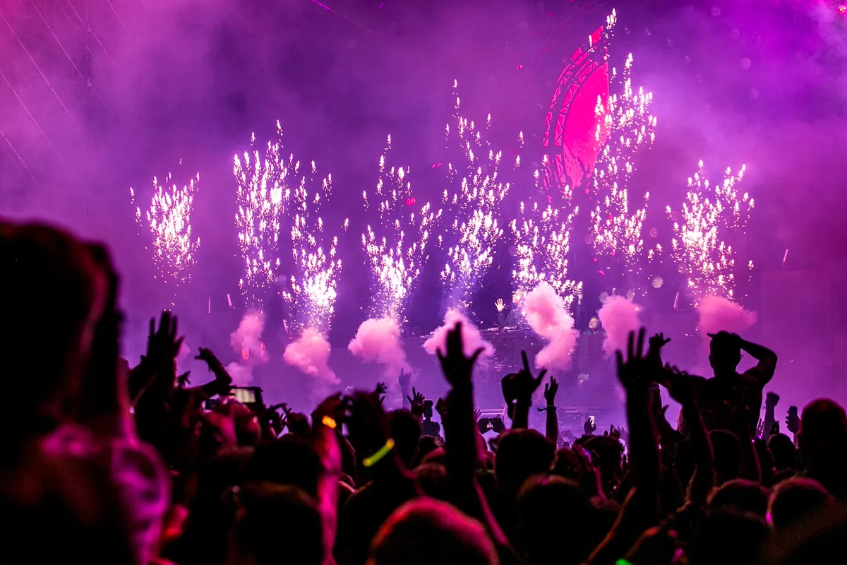 Foule en liesse aux mains levées lors d'un concert, éclairée par des étincelles pyrotechniques et une lumière violette intense.
