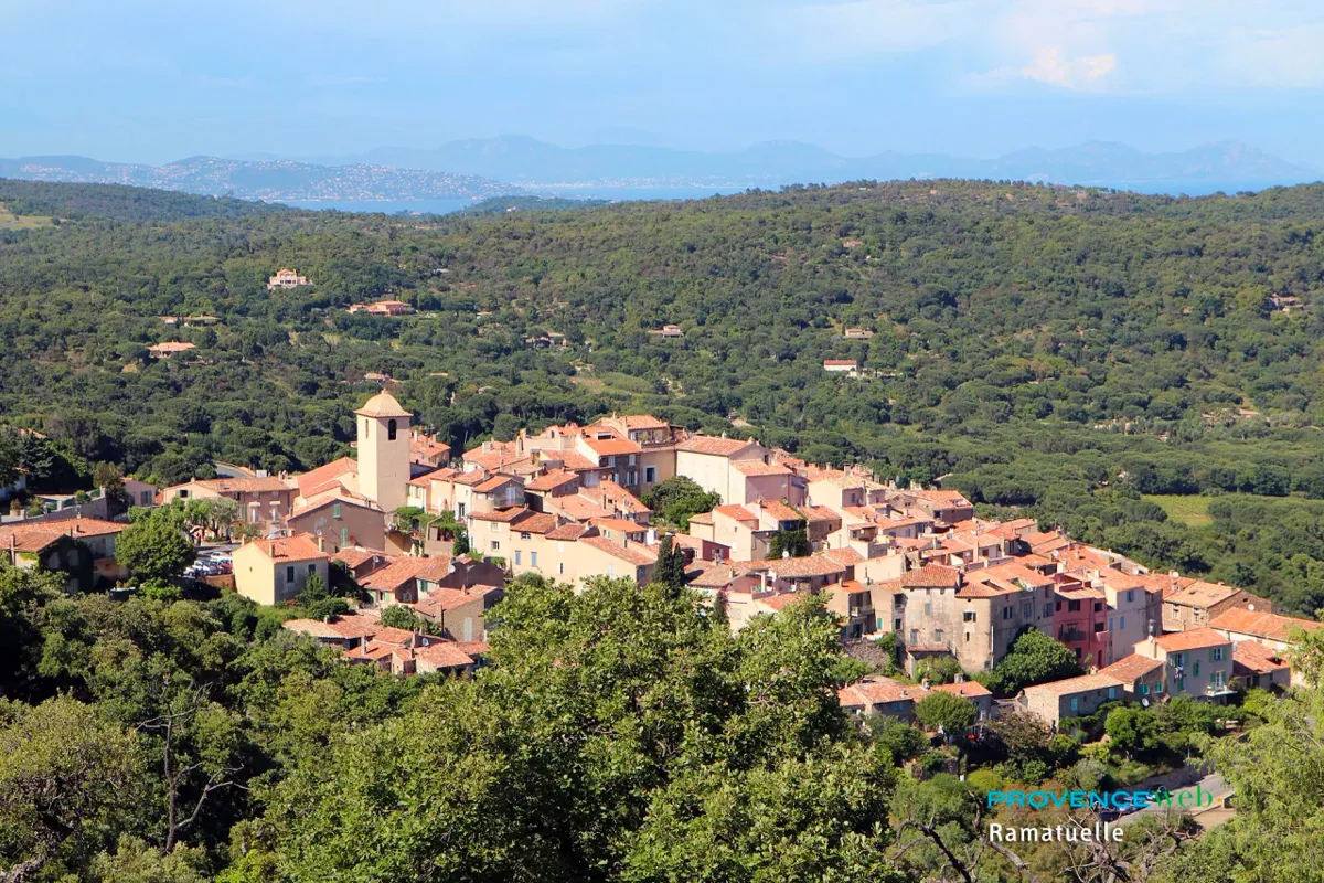 Vue aérienne du village perché de Ramatuelle en Provence, avec son clocher et ses toits en tuiles orange, entouré de collines boisées et d'un panorama sur la mer au loin.