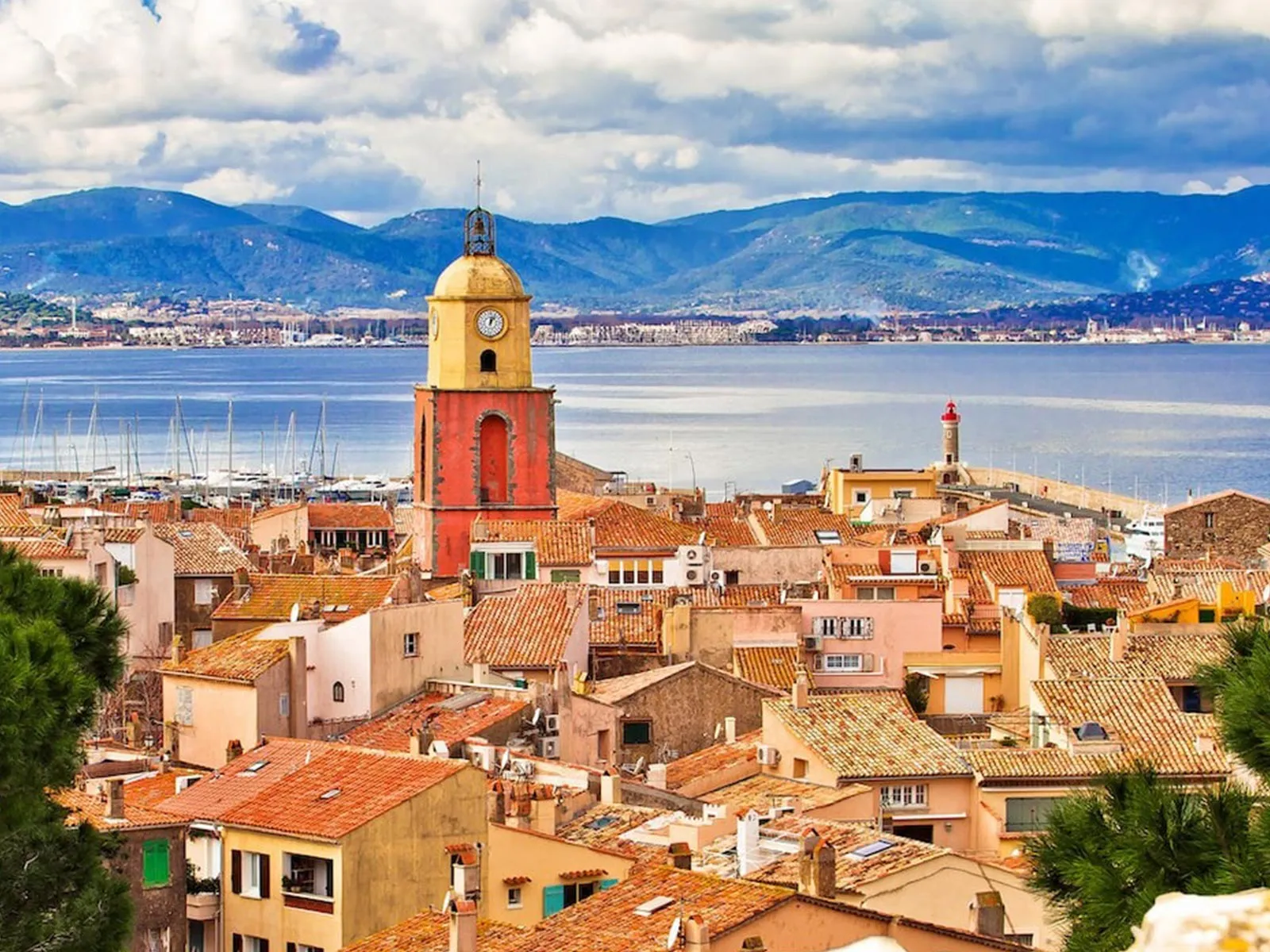Vue surélevée de Saint-Tropez dominée par son clocher rouge et jaune, entre toits de tuiles orange et la baie encadrée de montagnes.
