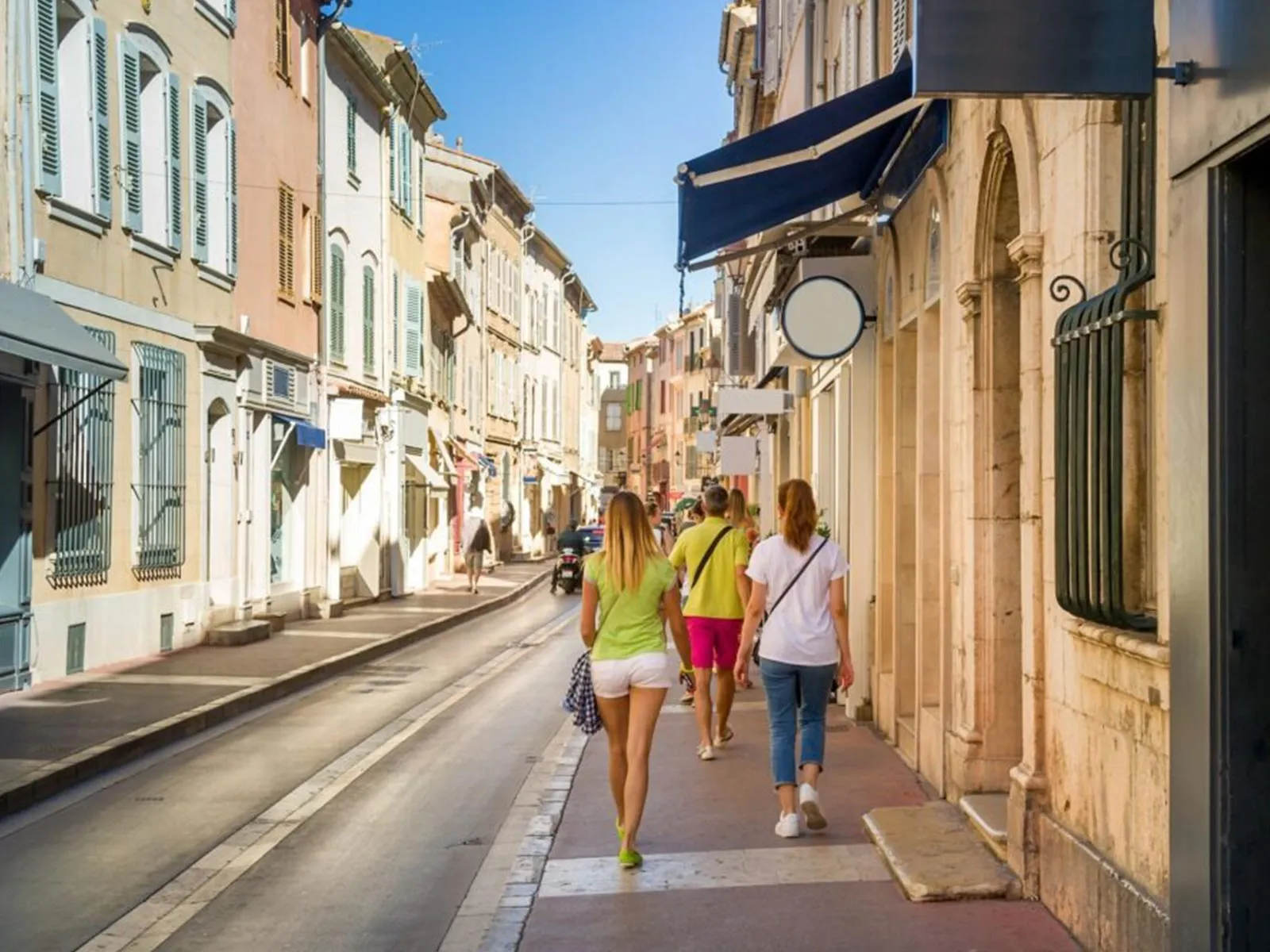 Rue commerçante ensoleillée bordée de bâtiments pastel aux volets colorés, avec des passants flânant devant des boutiques à Saint-Tropez.
