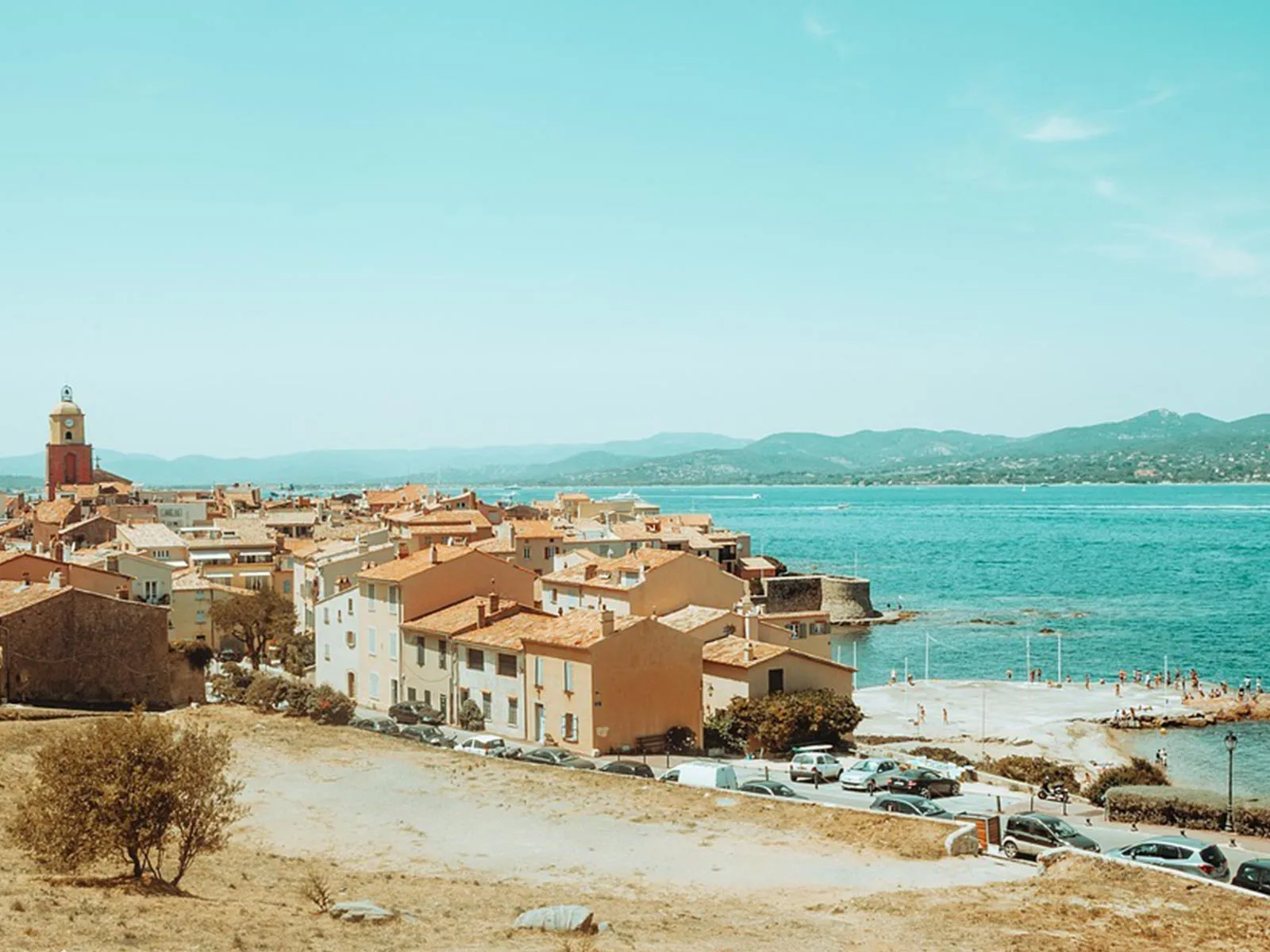 Vue panoramique de Saint-Tropez avec son clocher rouge, ses maisons aux toits de tuiles bordant la plage, et la mer turquoise s'étendant vers les collines.