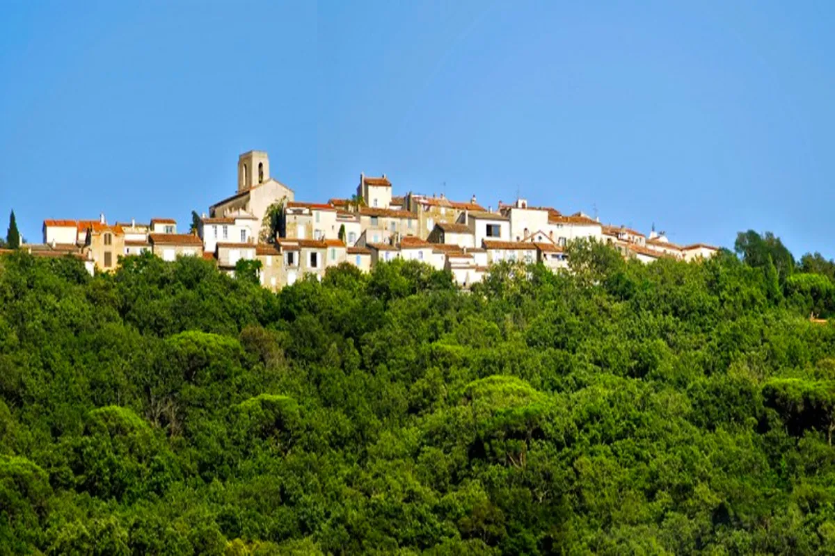 Village provençal perché avec son clocher et ses maisons aux toits de tuiles, émergeant d'une colline densément boisée sous un ciel bleu clair.