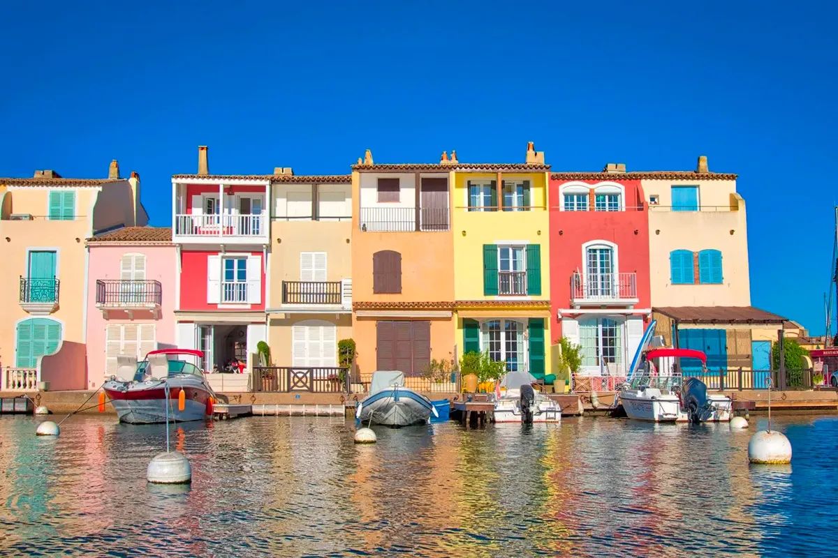 Rangée de maisons colorées aux volets bleus et vert menthe bordant un canal de Port Grimaud, avec bateaux amarrés sous un ciel bleu intense.