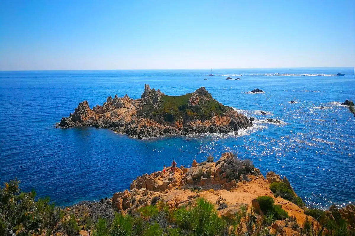 Îlot rocheux aux formations déchiquetées émergeant d'une mer méditerranéenne bleu intense, vu depuis une falaise ocre où se tiennent deux randonneurs.