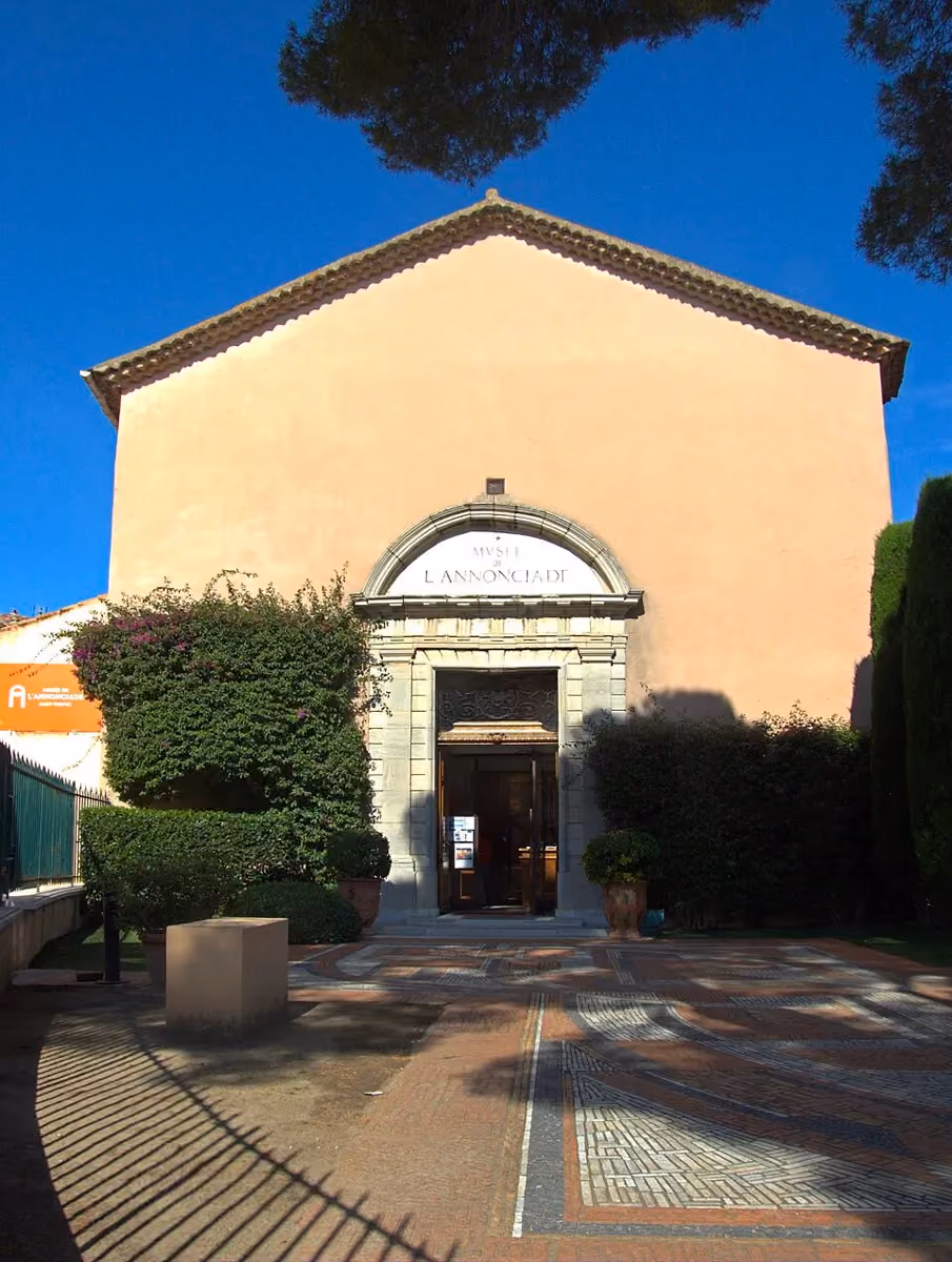 Façade du Musée de l'Annonciade à Saint-Tropez, ancienne chapelle aux murs rose pâle avec porche en pierre sculptée, entourée de buissons et d'un parvis en mosaïque.