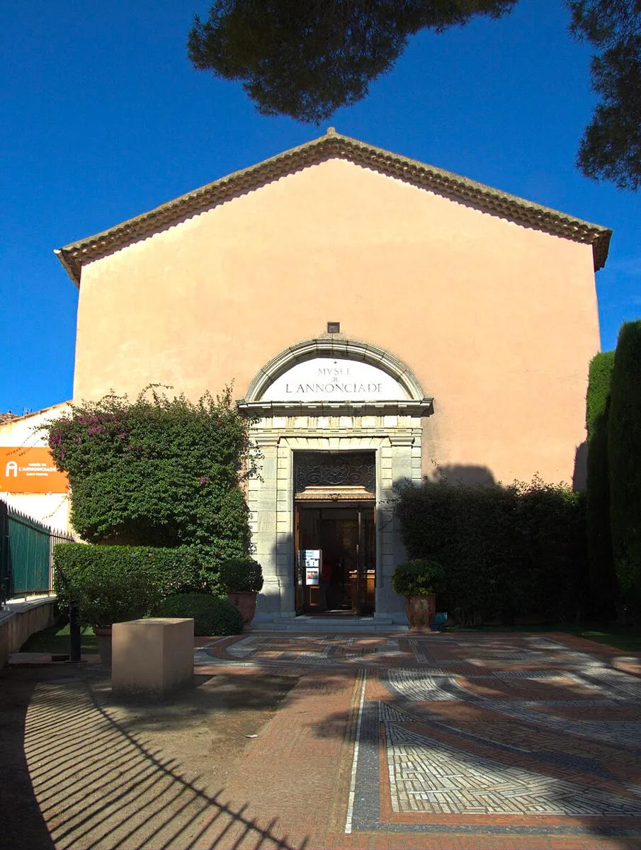 Façade du Musée de l'Annonciade à Saint-Tropez, ancienne chapelle aux murs rose pâle avec porche en pierre sculptée, entourée de buissons et d'un parvis en mosaïque.
