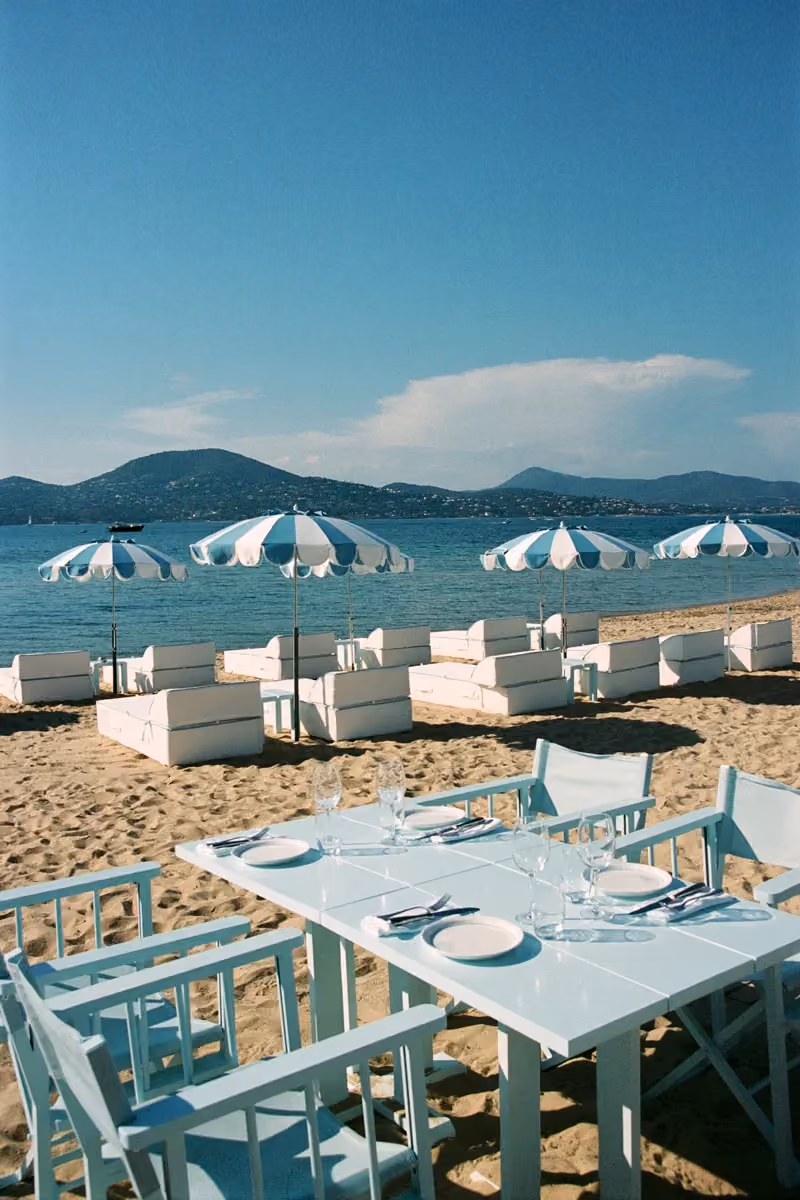 Restaurant de plage aux tables bleu pâle dressées, entouré de canapés et parasols rayés blanc et bleu sur le sable, avec vue sur la mer et les collines.