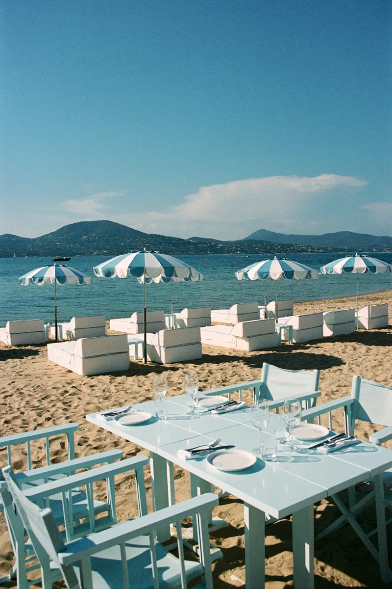 Restaurant de plage aux tables bleu pâle dressées, entouré de canapés et parasols rayés blanc et bleu sur le sable, avec vue sur la mer et les collines.