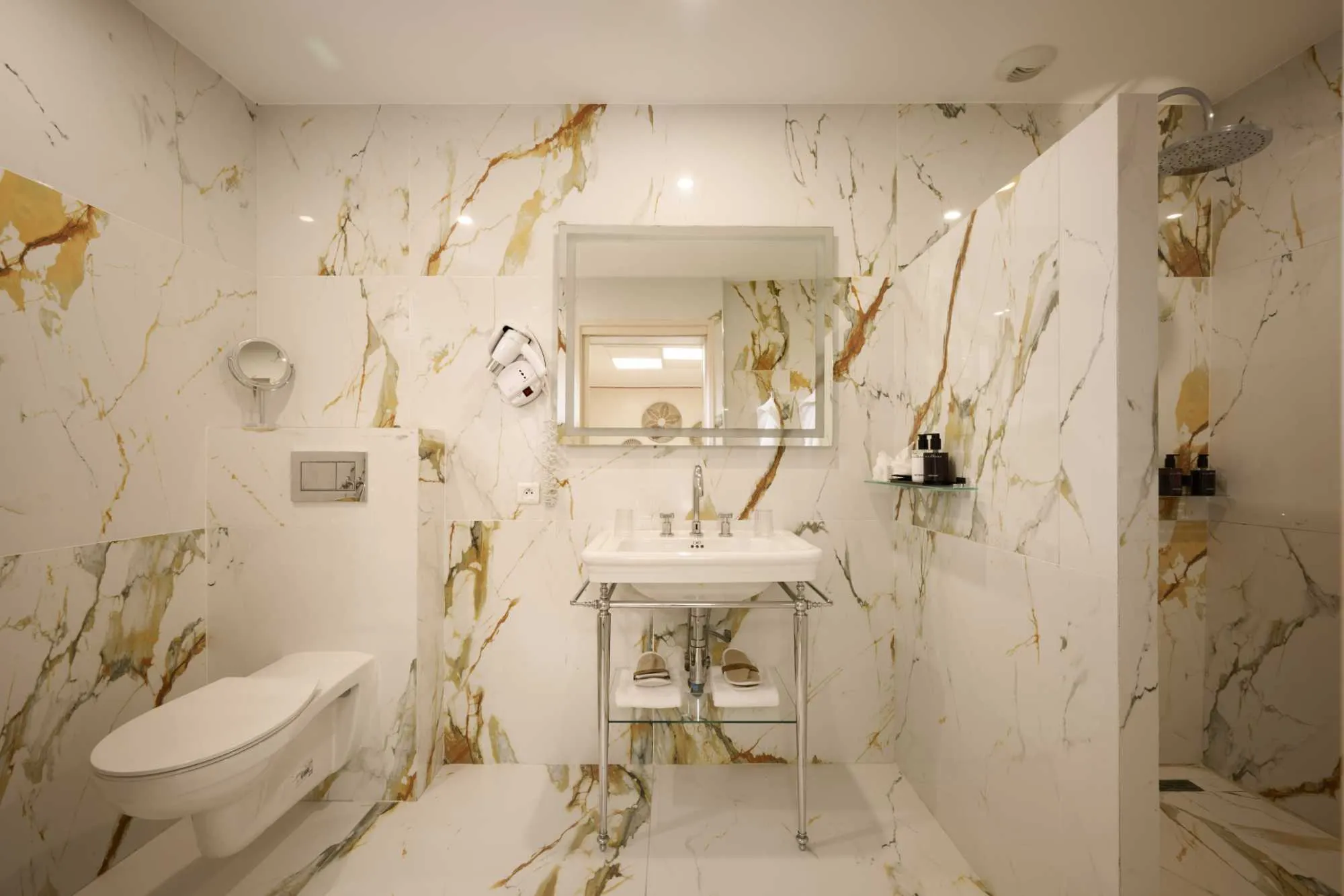Salle de bain luxueuse entièrement recouverte de marbre blanc veiné d'or, avec vasque sur pieds chromés, WC suspendu et douche ouverte.