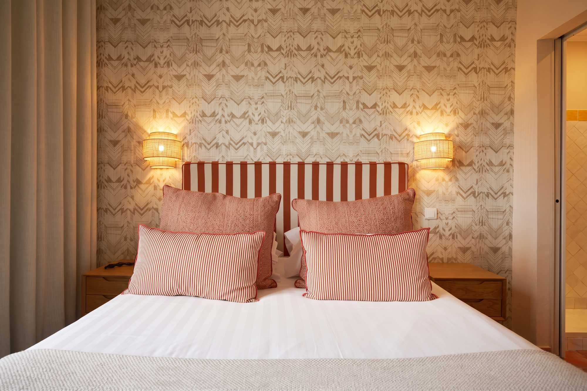 Gros plan sur un lit à tête rayée orange et blanc, garni de coussins rayés et texturés, encadré d'appliques tressées et de papier peint à motifs chevrons.