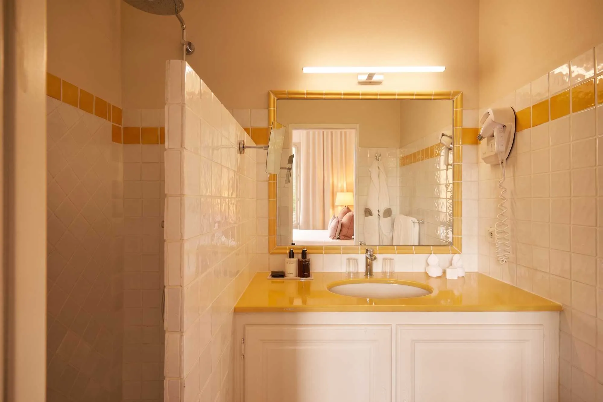 Salle de bain ensoleillée aux tons jaune et blanc, avec vasque et miroir bordés de carreaux jaunes, meuble blanc, et douche ouverte carrelée en losange.
