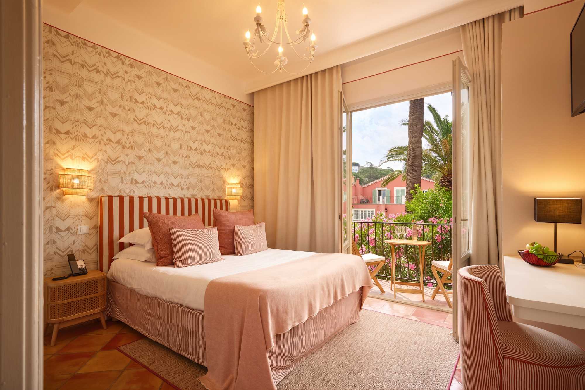 Chambre romantique aux tons rose poudré avec lustre orné, lit rayé, et balcon fleuri offrant une vue sur des palmiers et une villa rose.