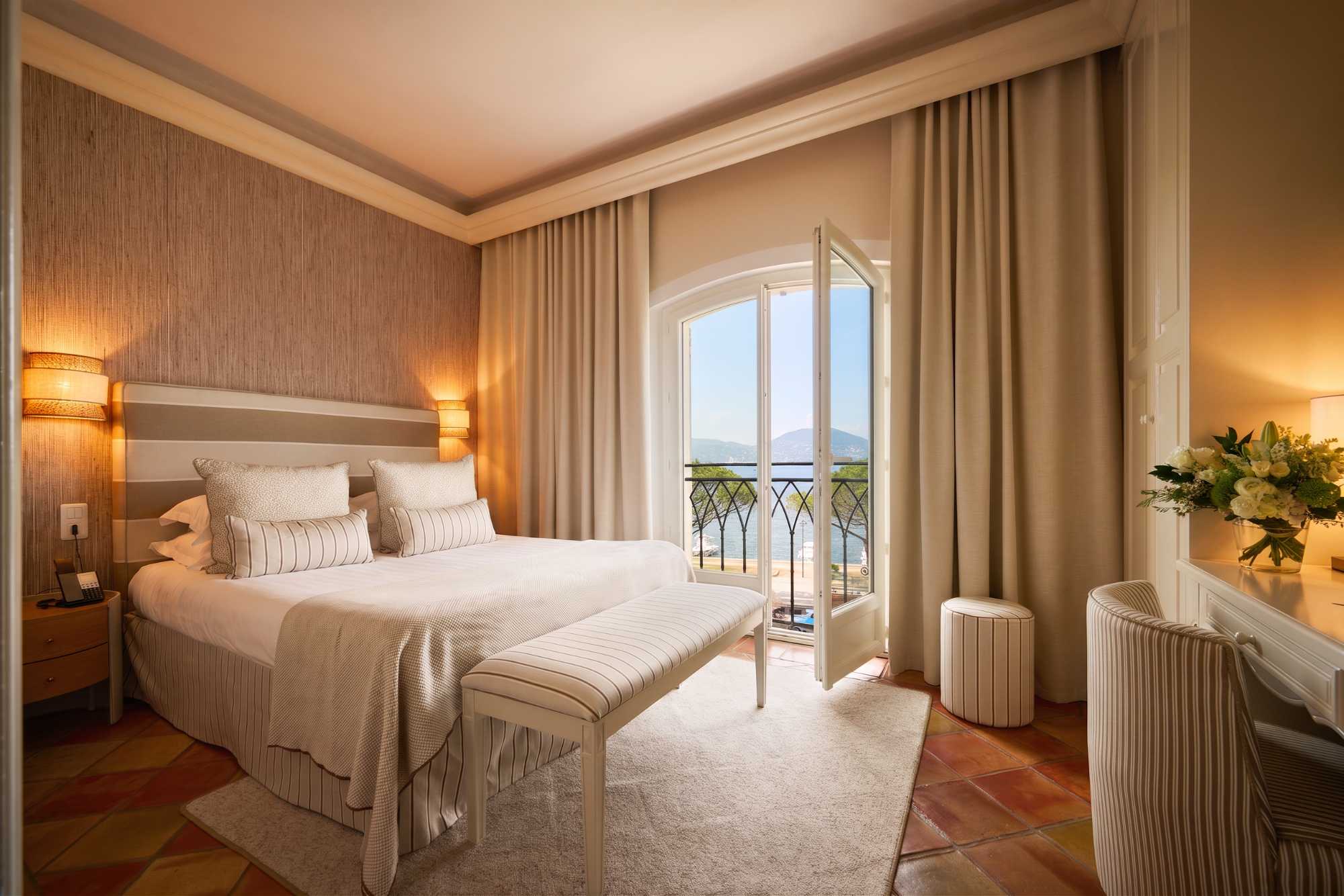 Chambre élégante aux tons beige avec banquette rayée, sol en terre cuite, et porte-fenêtre ouverte sur un balcon en fer forgé offrant une vue sur la mer et les collines.