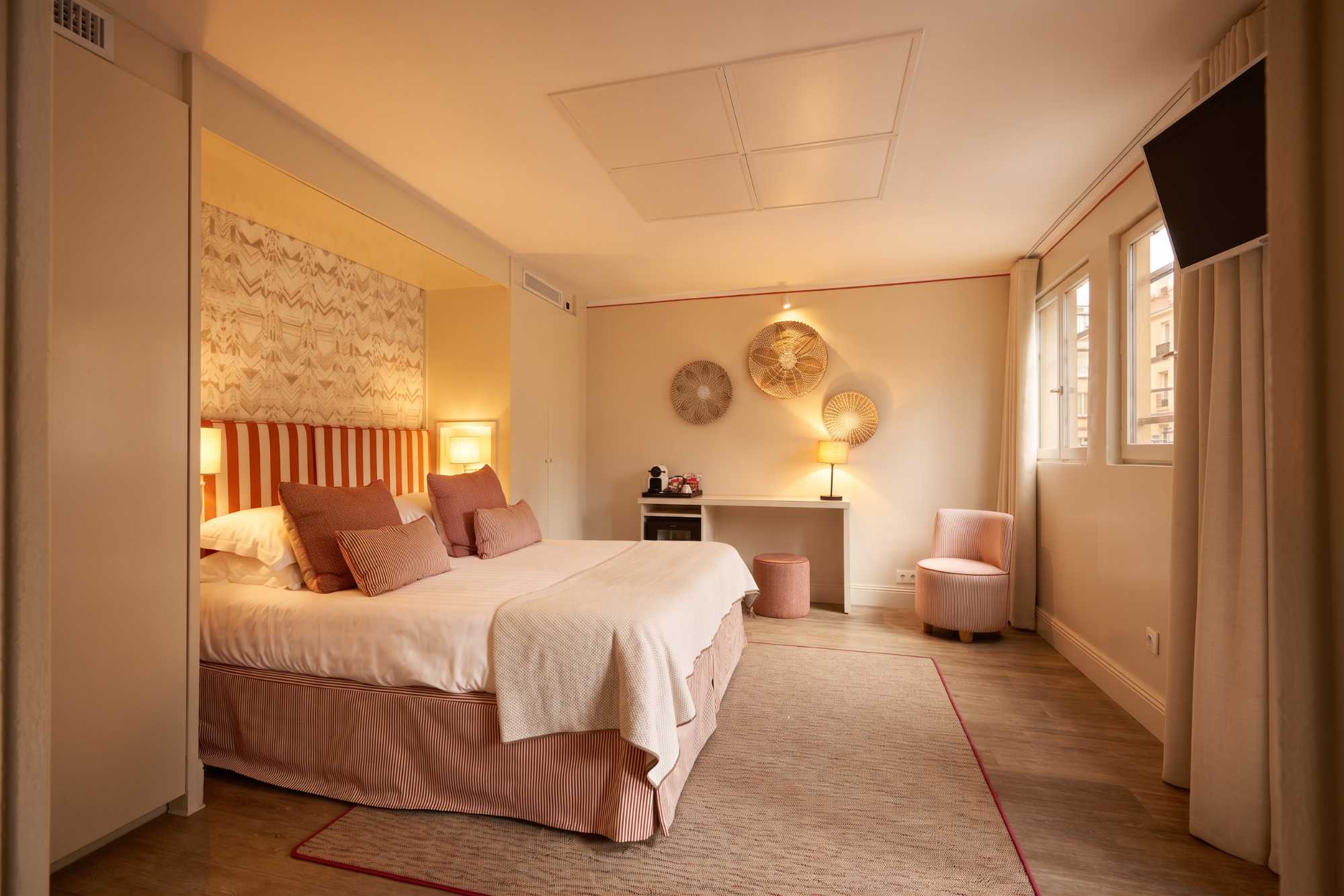Chambre spacieuse aux tons pêche et corail, avec lit rayé en alcôve, décorations murales en vannerie, bureau blanc et fauteuil assorti rayé.