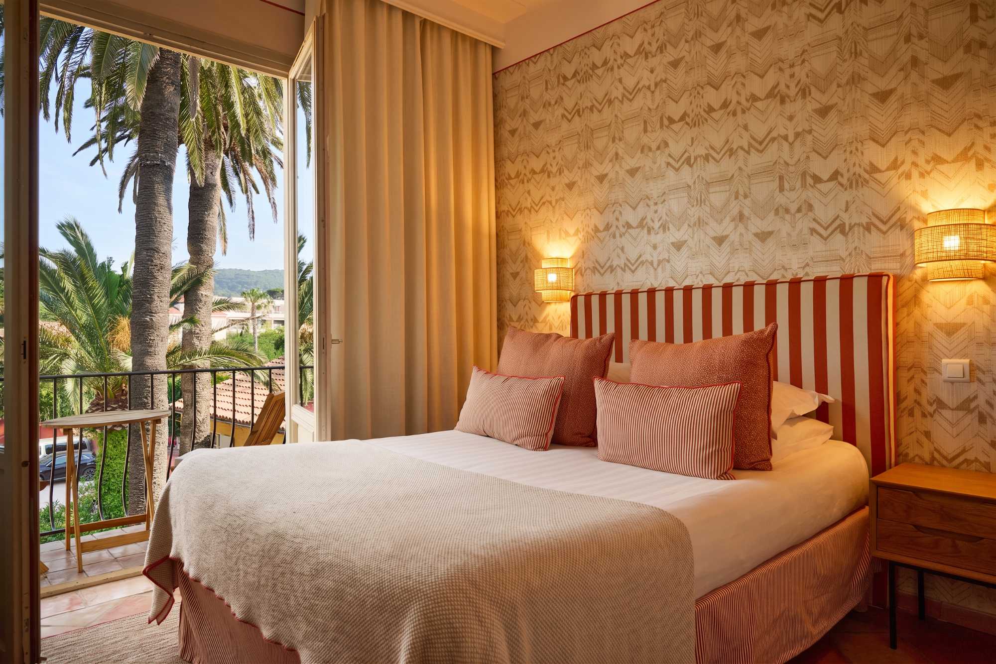 Chambre chaleureuse au papier peint à motifs et tête de lit rayée orange, avec balcon ouvert offrant une vue sur de hauts palmiers et les collines environnantes.