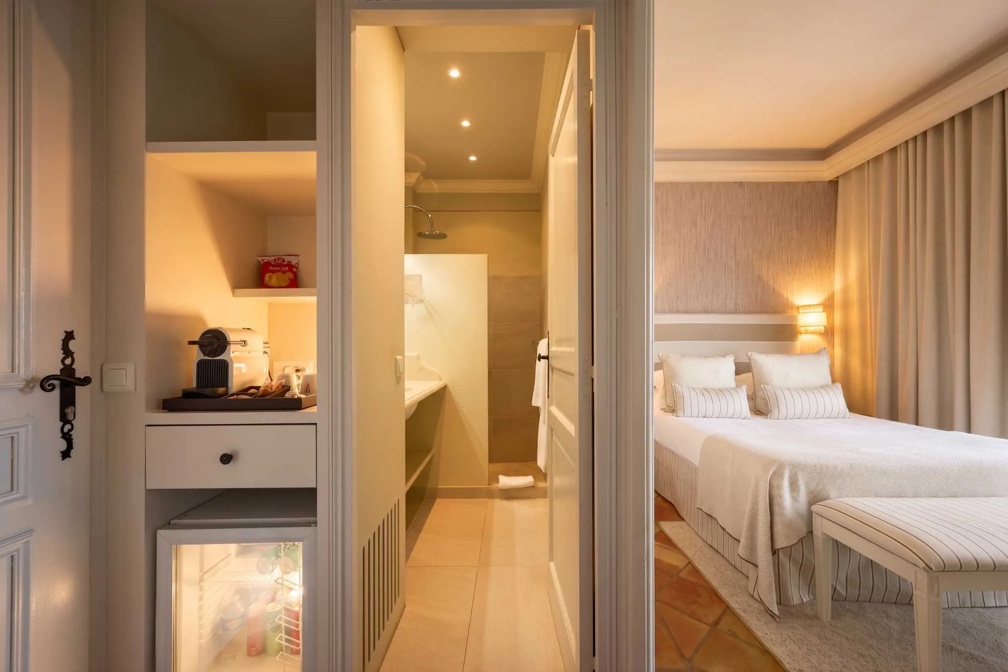 Vue composite d'une chambre d'hôtel montrant un placard avec minibar et machine à café, une salle de bain attenante, et un lit habillé de rayures beige.
