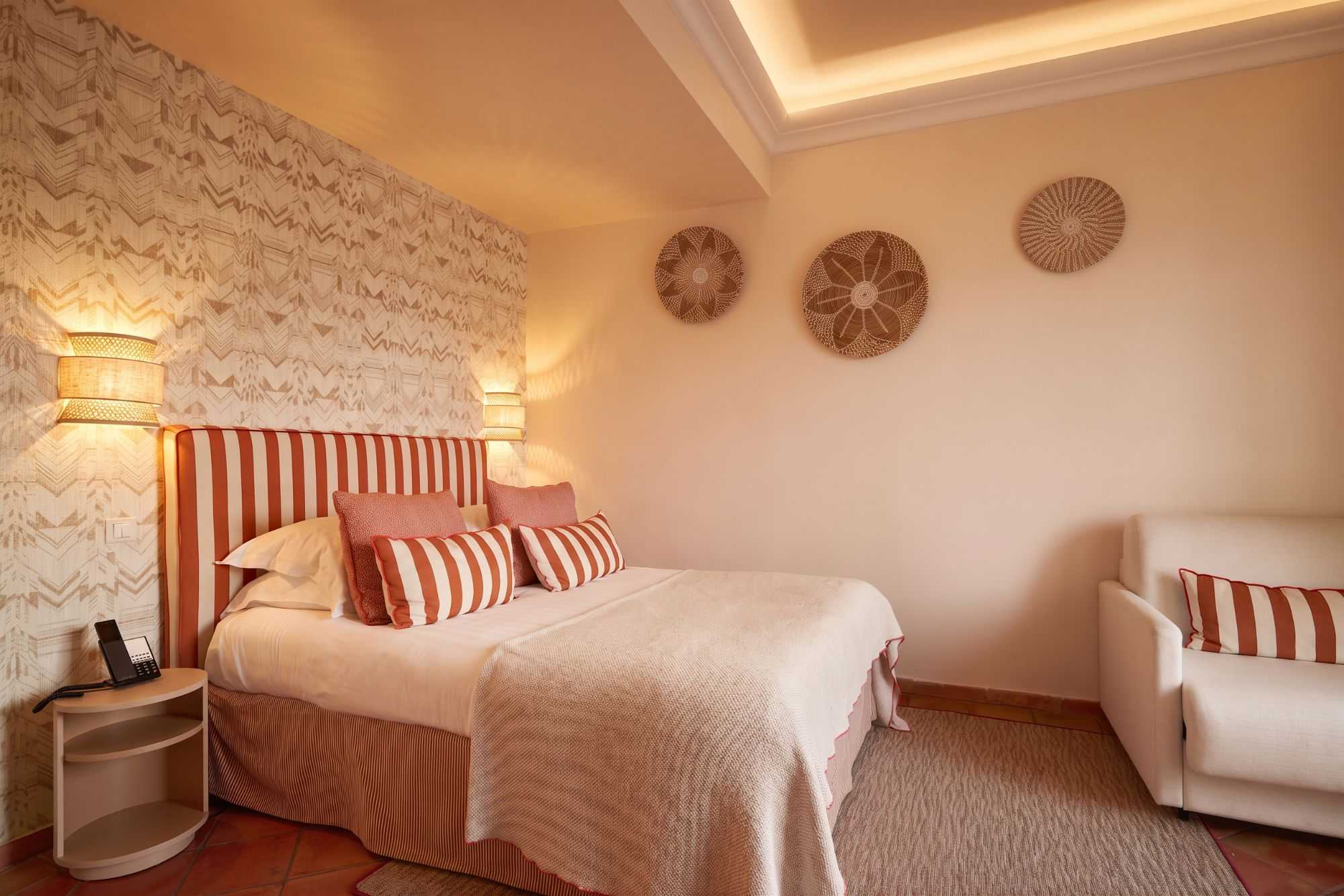 Chambre confortable aux tons corail, avec lit et fauteuil rayés, papier peint à motifs, décorations murales en vannerie, et plafond à corniche éclairée.