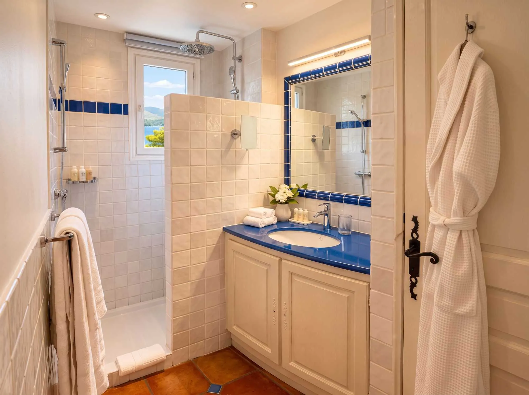 Salle de bain provençale carrelée en blanc et bleu, avec vasque à plan bleu vif, douche ouverte avec vue sur la mer par la fenêtre, et peignoir suspendu à la porte.
