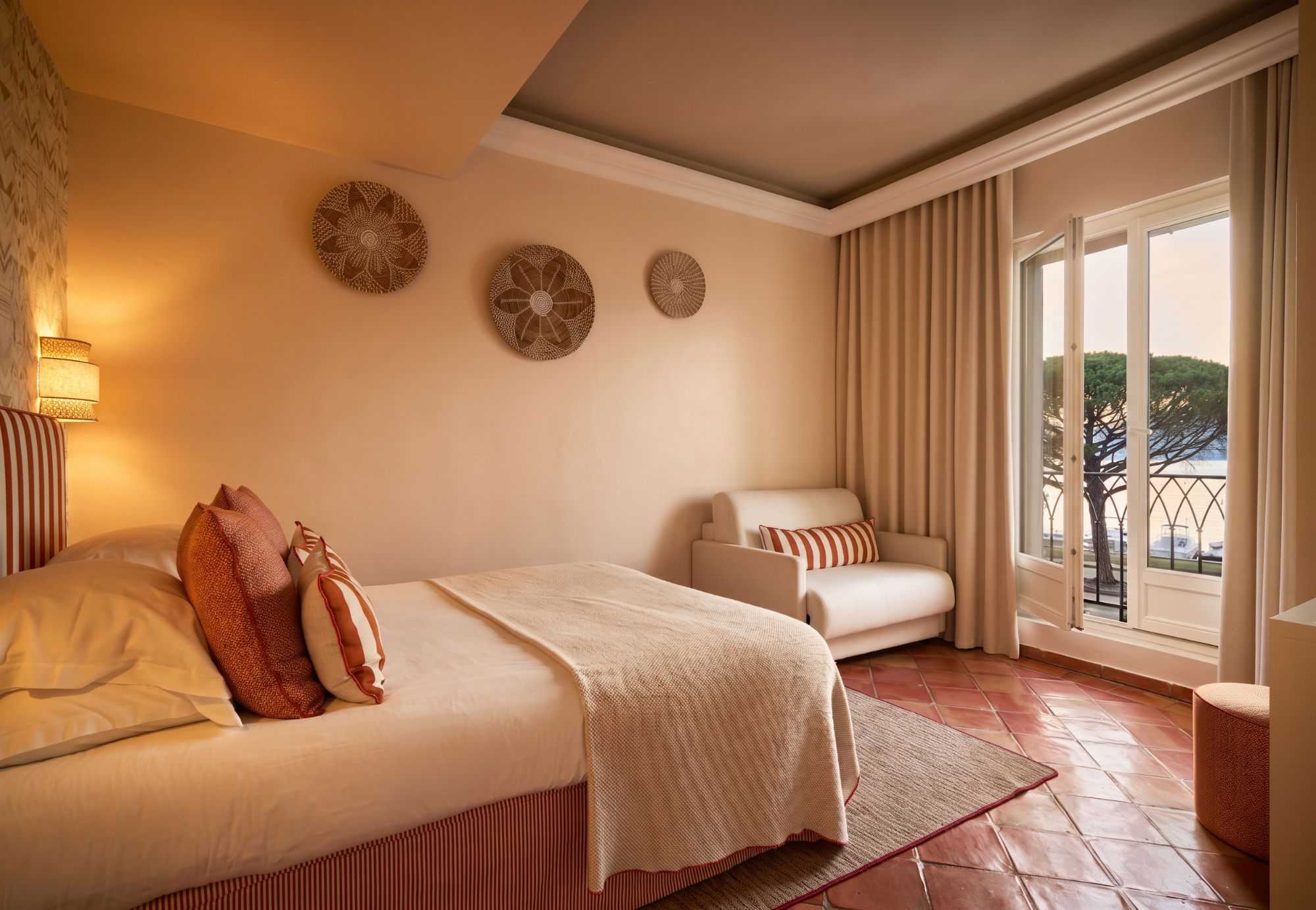 Chambre chaleureuse aux tons terracotta, décorée de vanneries murales, avec lit et fauteuil crème, sol en tomettes, et balcon en fer forgé donnant sur des pins et la mer.