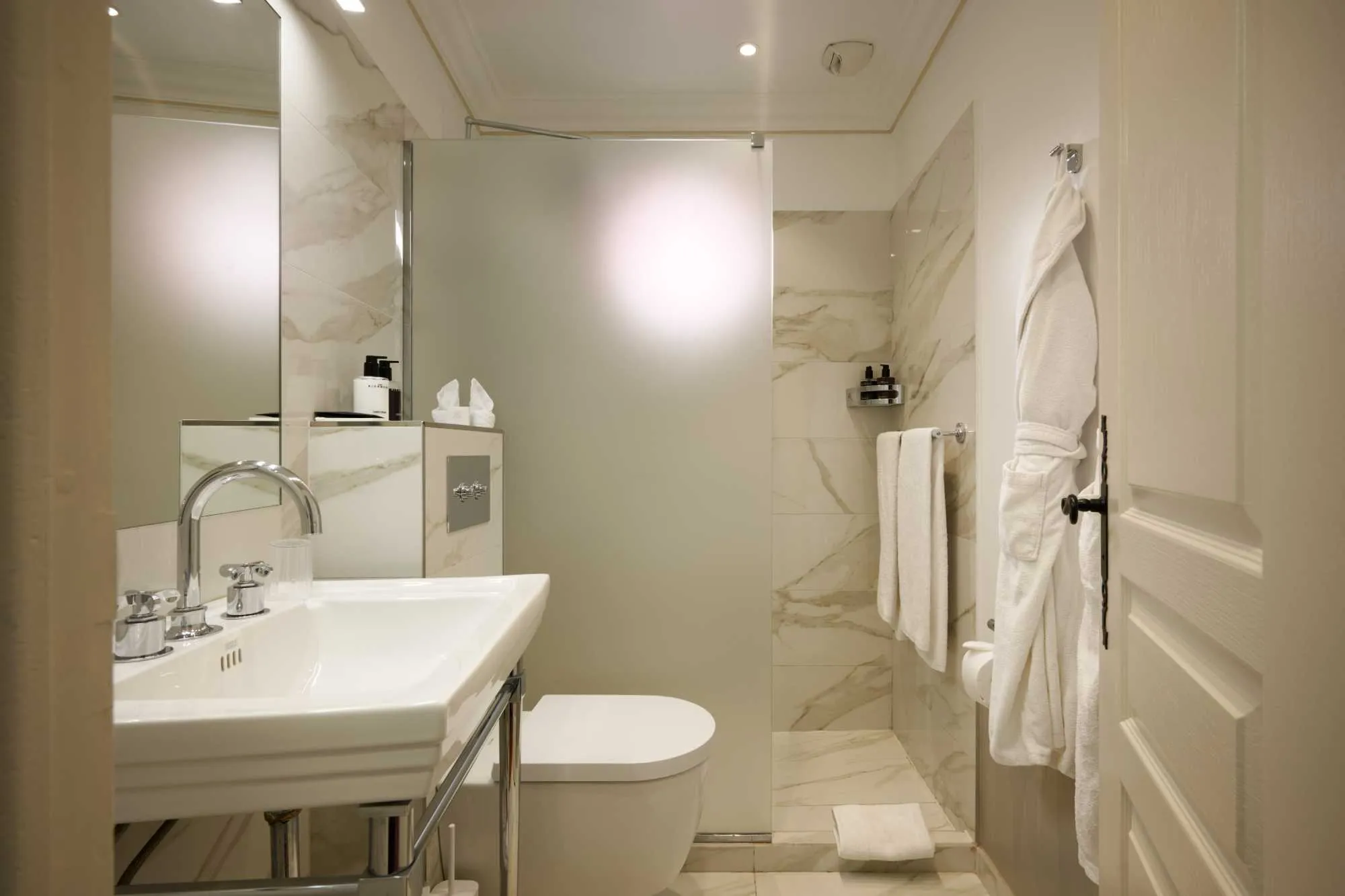 Salle de bain élégante en marbre beige veiné, avec vasque sur pieds chromés, douche vitrée dépolie, WC, et peignoir blanc suspendu à la porte.