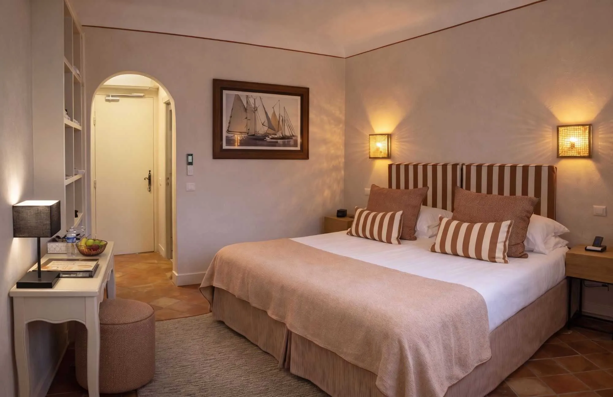 Chambre chaleureuse aux tons camel, avec lit rayé, coiffeuse blanche, tableau de voiliers, entrée voûtée et sol en terre cuite typique provençal.