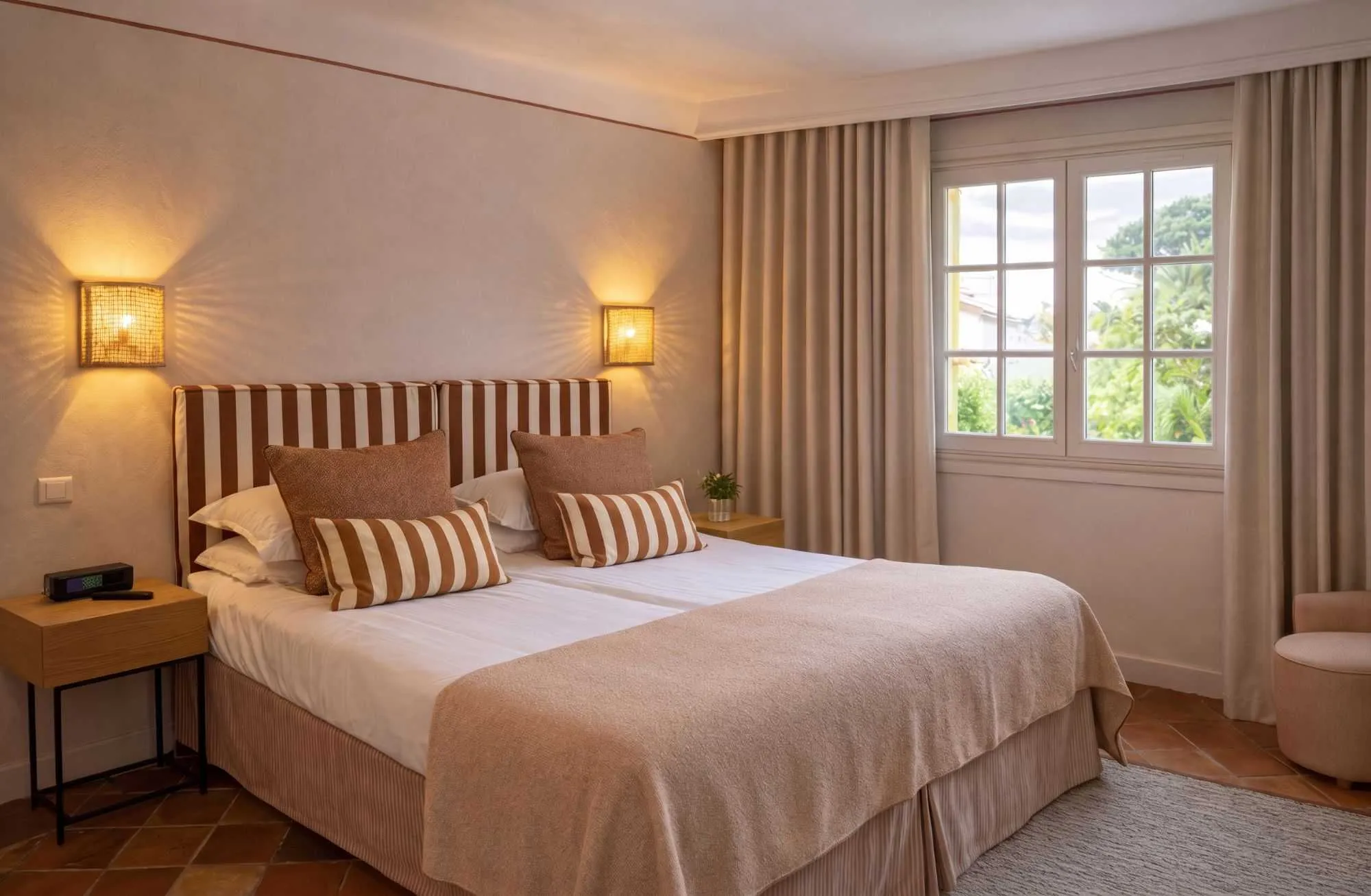 Chambre douillette aux tons camel et beige, avec tête de lit et coussins rayés, appliques lumineuses tressées, sol en terre cuite et fenêtre donnant sur la végétation.