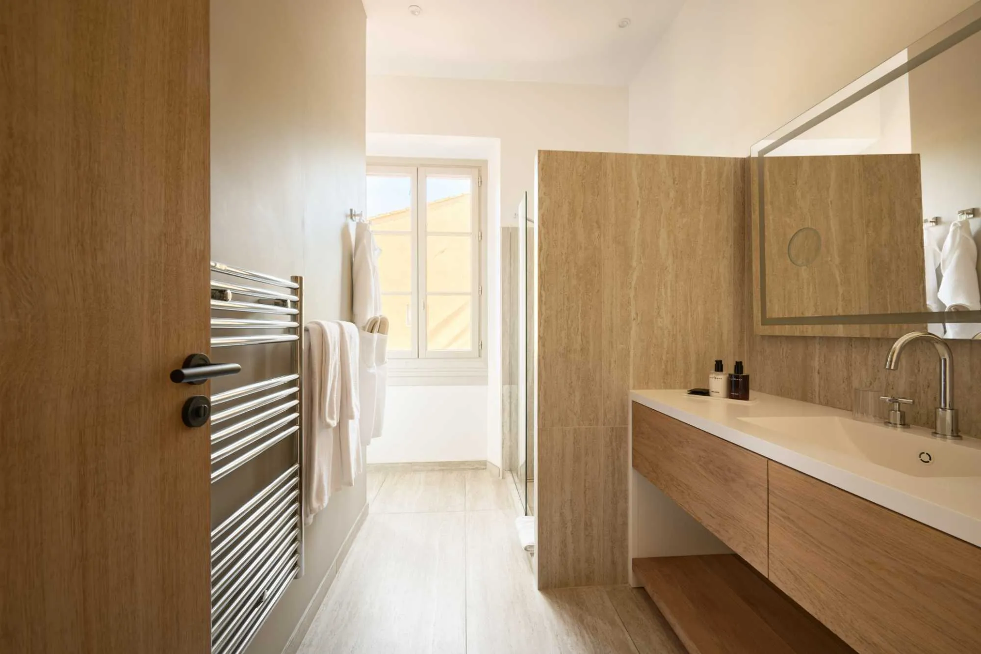 Salle de bain claire avec porte en bois ouverte, sèche-serviettes chromé, vasque en marbre travertin et miroir rétroéclairé, avec fenêtre au fond laissant entrer la lumière.