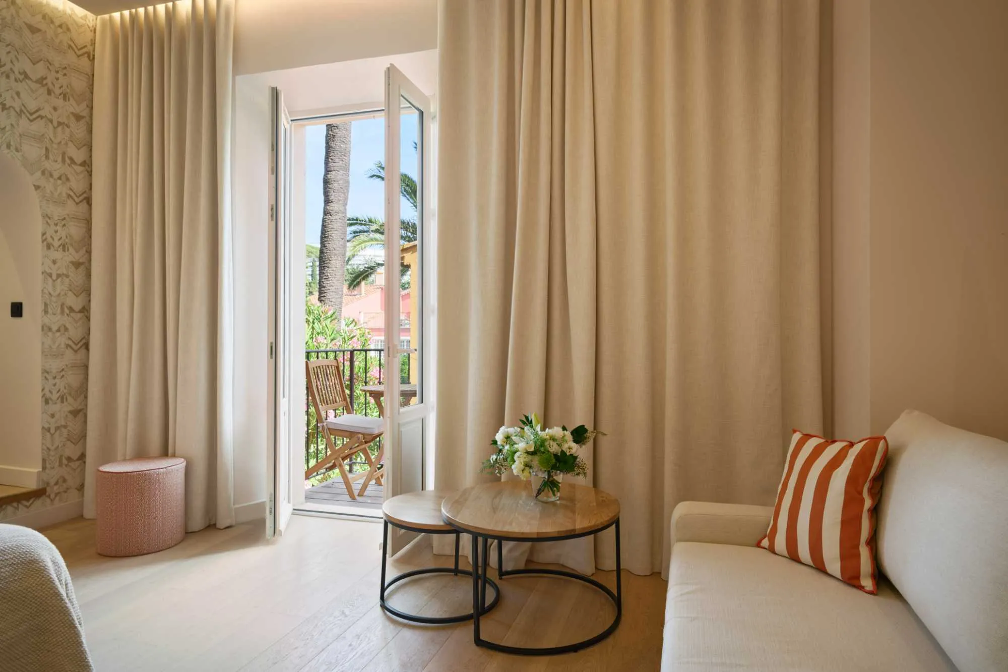 Coin salon aux tons neutres avec canapé et coussin rayé orange, tables basses gigognes fleuries, et rideaux beige encadrant une porte-fenêtre ouverte sur un balcon.
