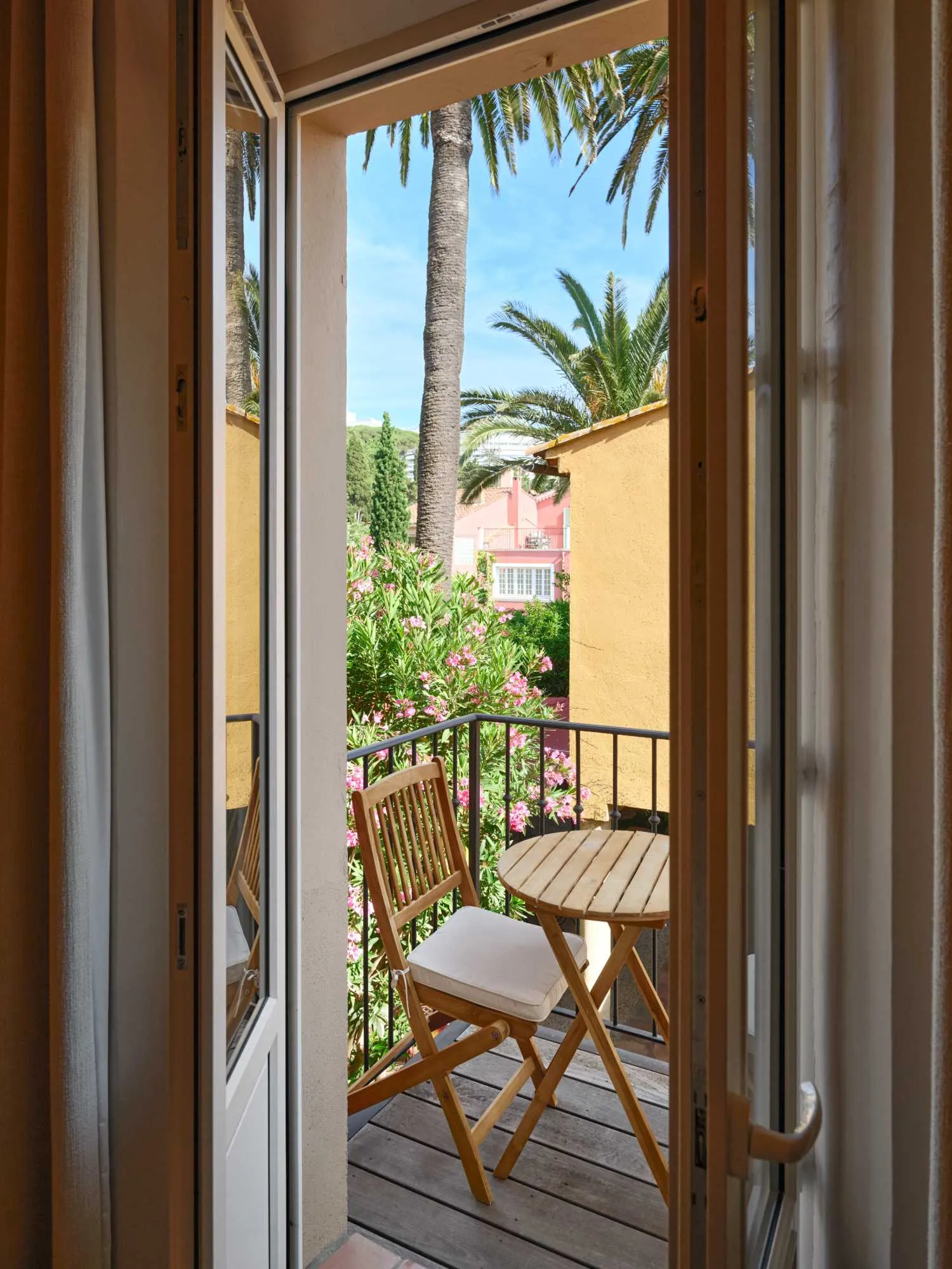 Vue depuis une porte-fenêtre ouverte sur un petit balcon en bois avec table et chaise, entouré de lauriers-roses et surplombé par un haut palmier et des façades ocre.