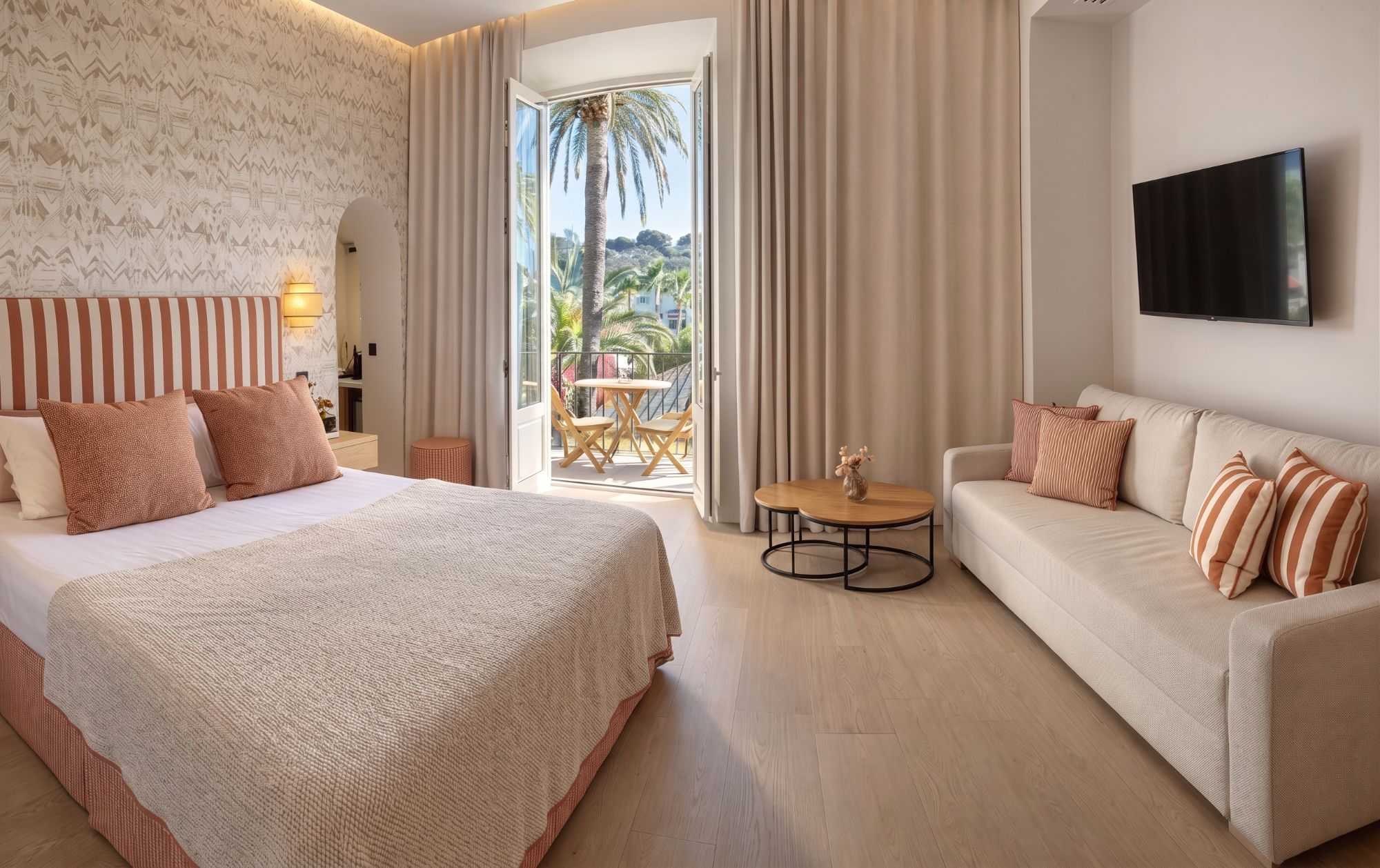 Suite spacieuse aux tons terracotta et beige, avec lit rayé, canapé assorti, télévision murale, et balcon ouvrant sur un grand palmier.